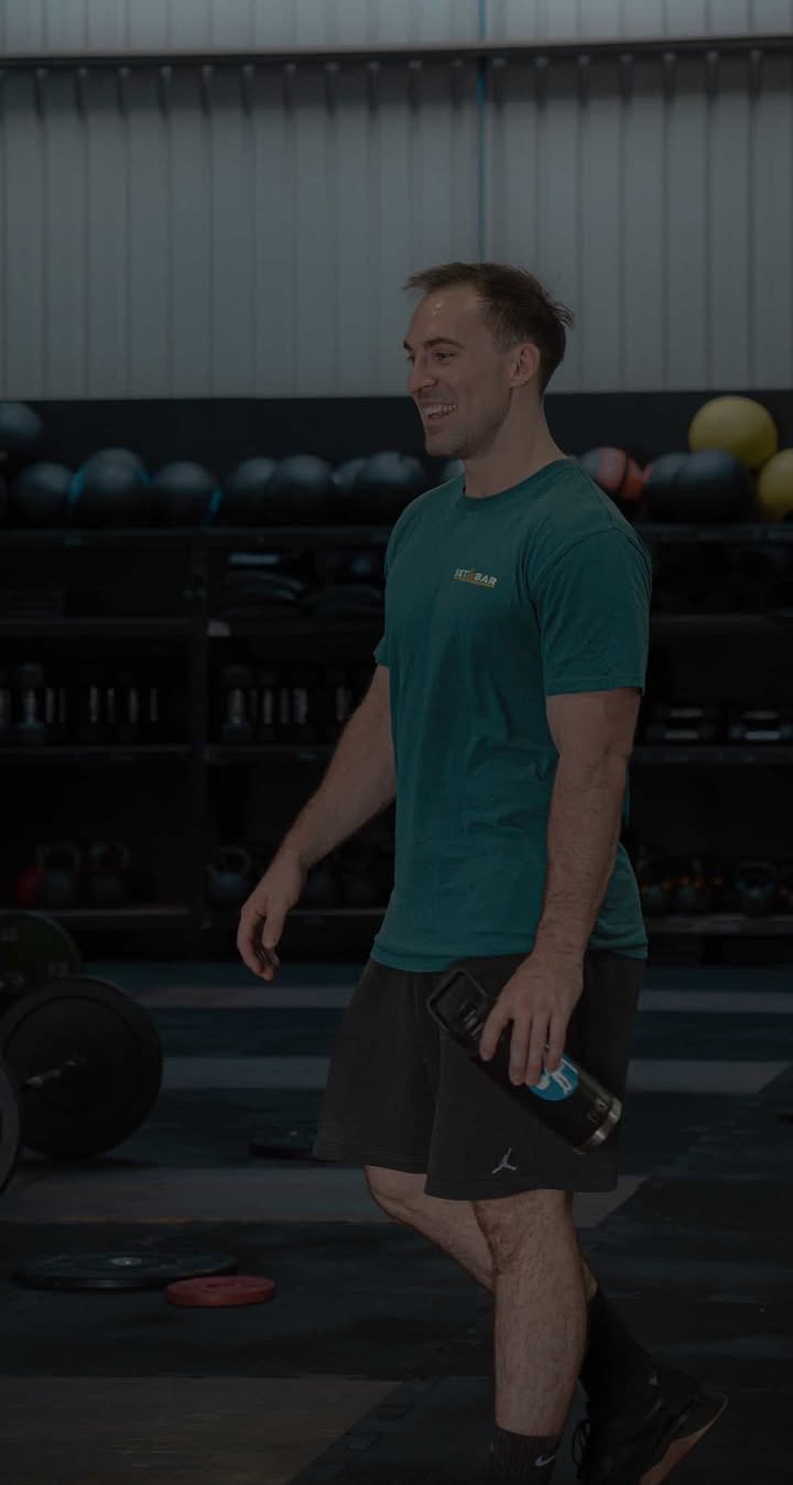A smiling young man holding a water bottle in a gym with weightlifting equipment in the background.