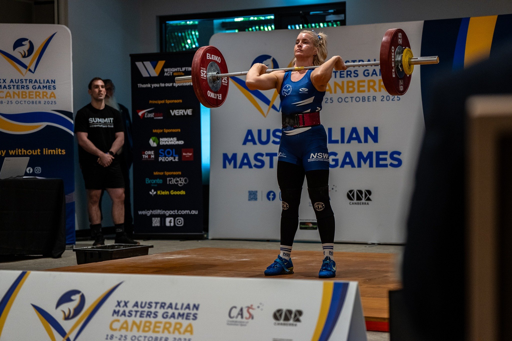 Female weightlifter in blue uniform lifting a barbell overhead during the Australian Masters Games in Canberra, October 2025, with officials and banners in the background.