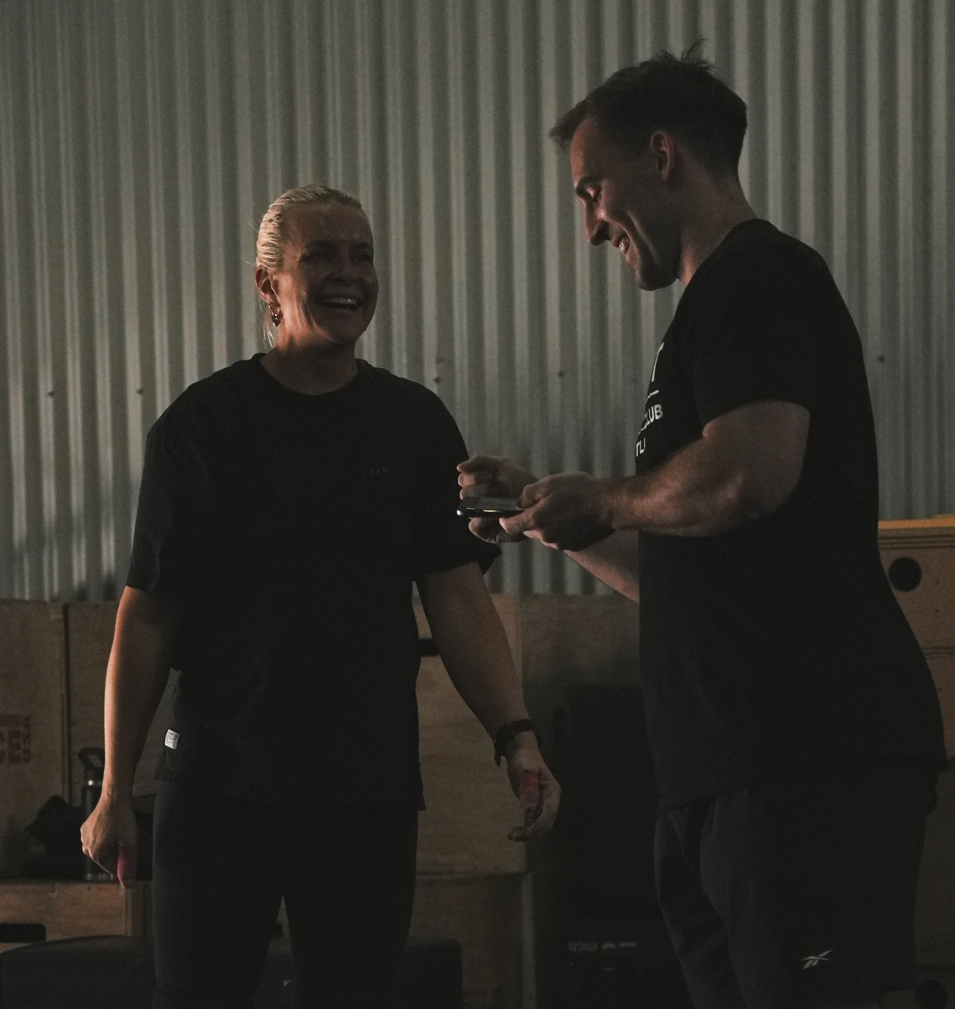 Two people in black athletic clothing smiling and interacting indoors against a corrugated metal wall.