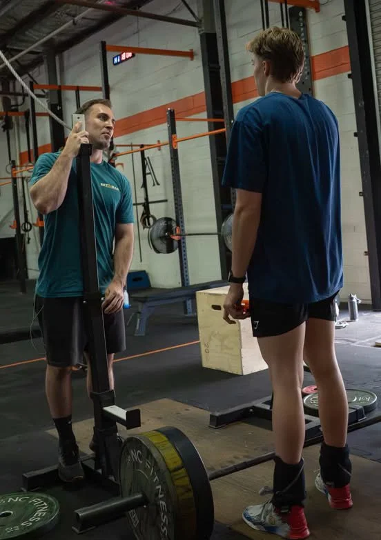 Two women in a gym, one taking a photo of the other who is preparing to lift a barbell with weights on a platform.