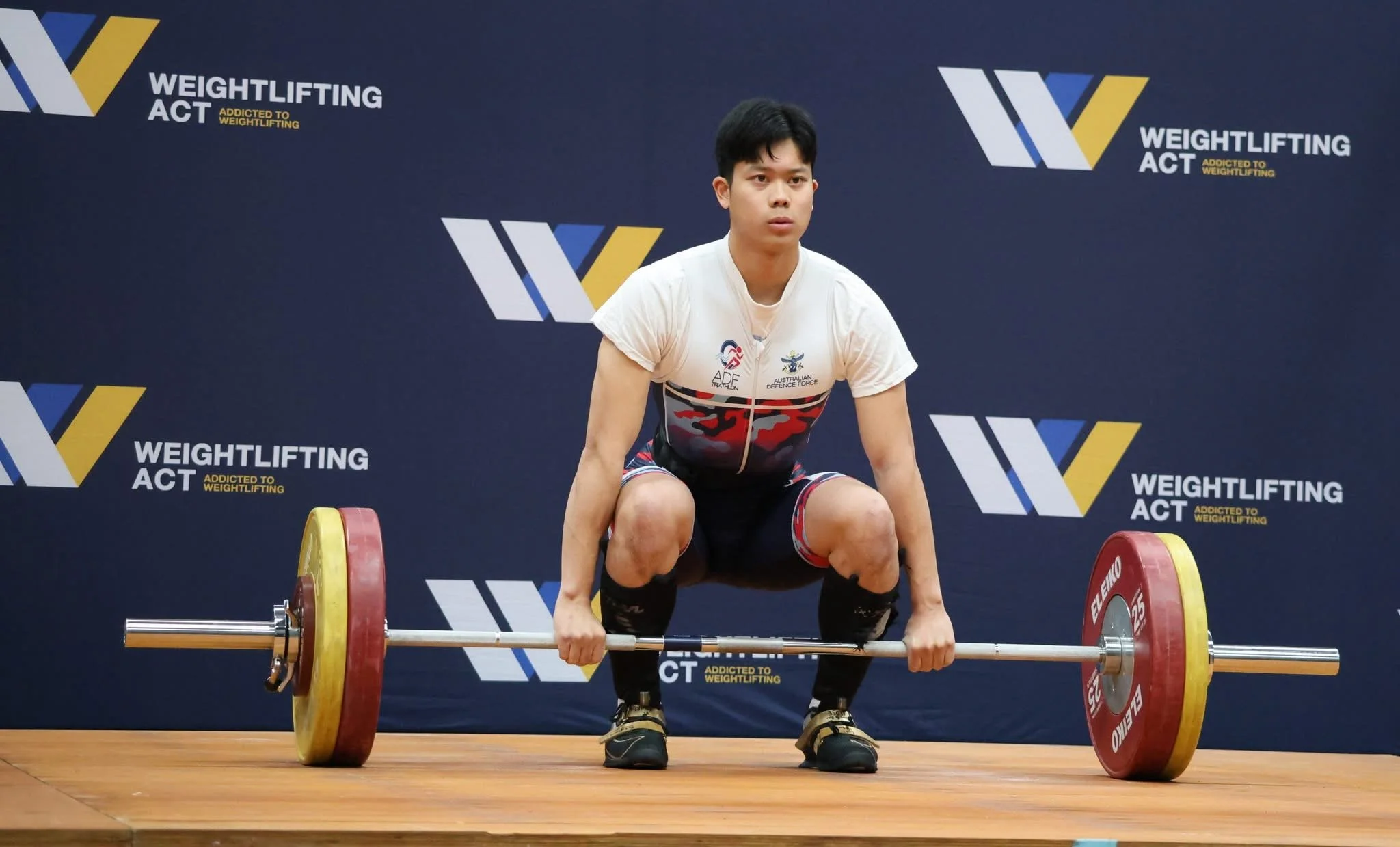 A young male weightlifter is preparing for a lift on a wooden platform in front of a dark backdrop with logos and text related to weightlifting, including "WEIGHTLIFTING ACT" and "ADDICTED TO WEIGHTLIFTING." He is squatting with a loaded barbell on the floor, wearing a white t-shirt with various logos, black shorts with red, white, and blue accents, and black weightlifting shoes.