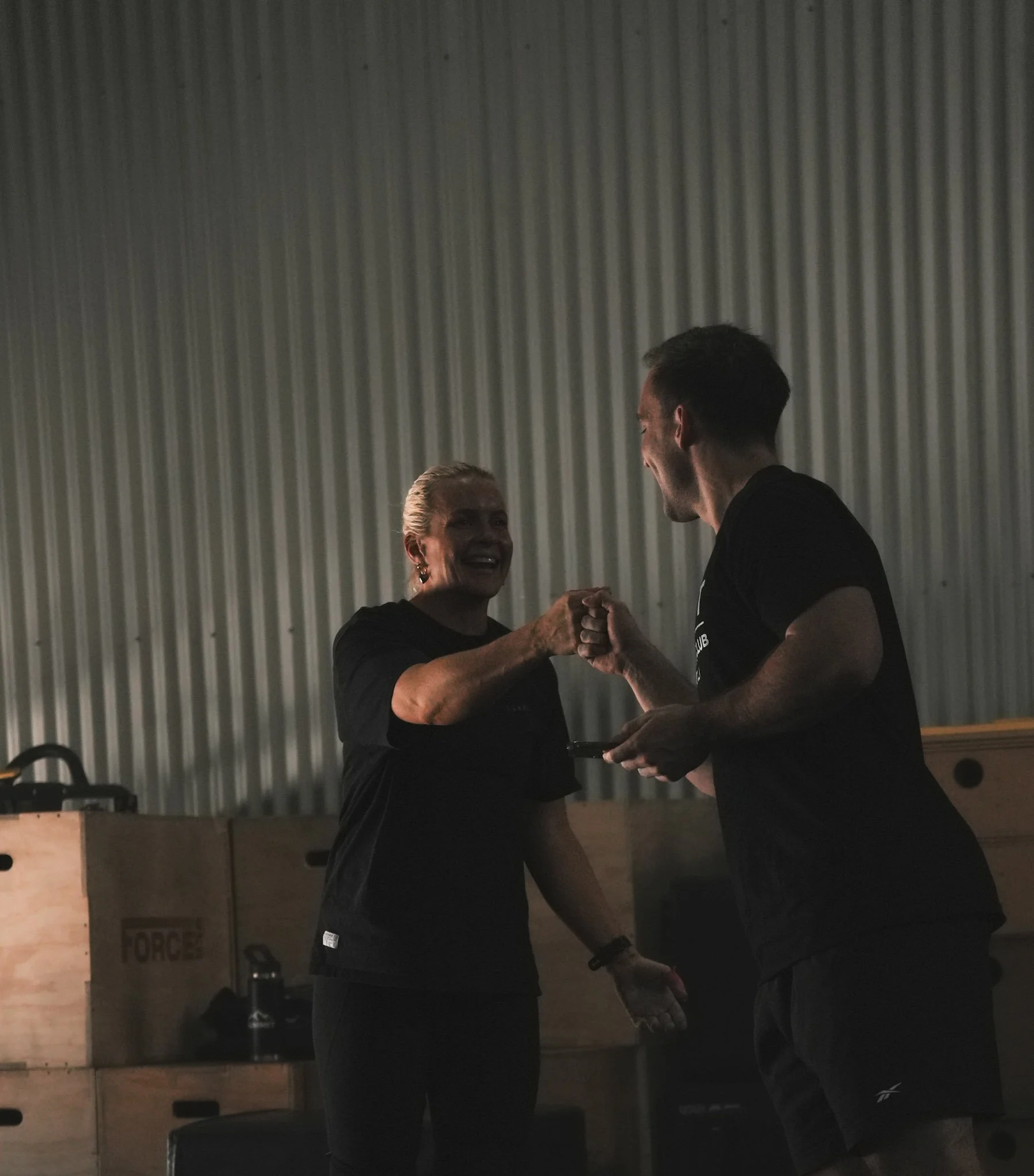 Two people, a woman and a man, smiling and shaking hands in a gym or fitness center, with wooden boxes and equipment in the background.