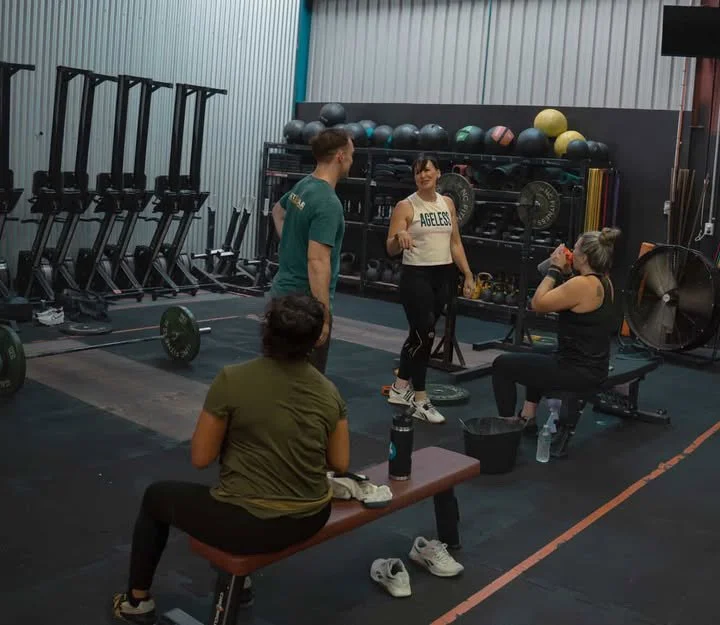 Four women in a gym, one standing and three seated, engaging in conversation and taking a break after workout, with various gym equipment in the background.