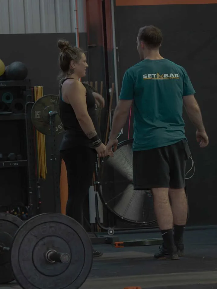 A woman and a man having a conversation in a gym, with gym equipment such as weights and a fan in the background.