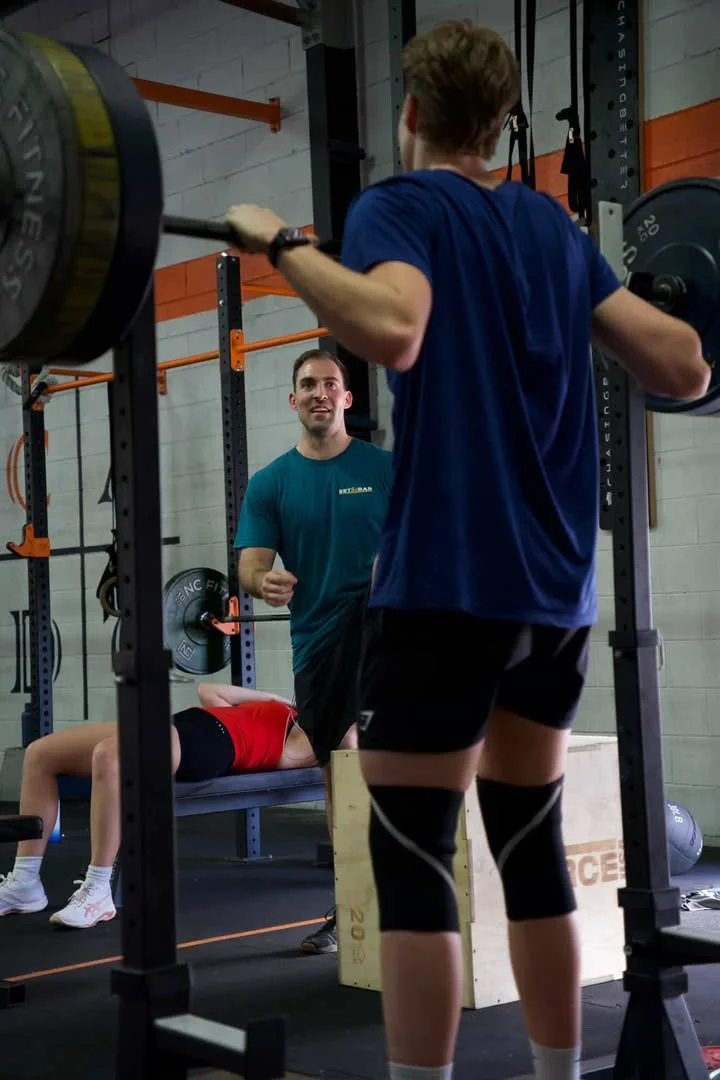 A woman in a red sports bra and black shorts performs a bench press at a gym. A man in a teal shirt watches her, smiling, as a young man in a blue shirt and black shorts stands nearby with a barbell on his shoulders, preparing to lift.