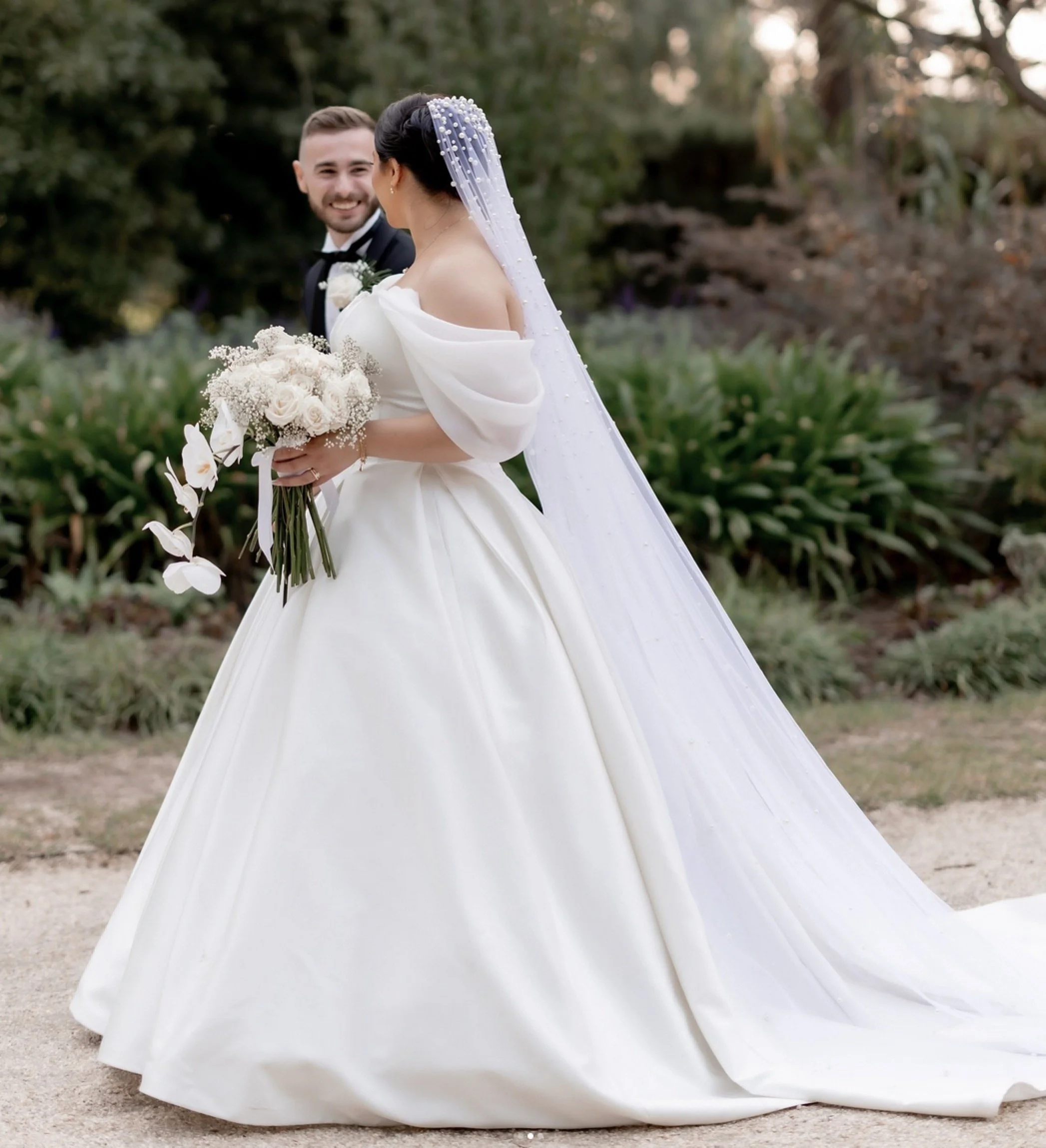 Bride and groom smiling at each other outdoors. The bride wears a white wedding gown with puffed sleeves and holds a bouquet of white roses and other flowers. The groom wears a black tuxedo with a bow tie. The bride's long veil has pearls, and they are standing on a dirt path with greenery and trees in the background.