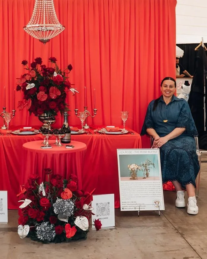 A woman sitting beside a decorated table with a large floral arrangement, set with glassware and plates, in front of a coral-colored curtain backdrop. There is a framed informational sign and floral decor on the floor with roses and butterflies.