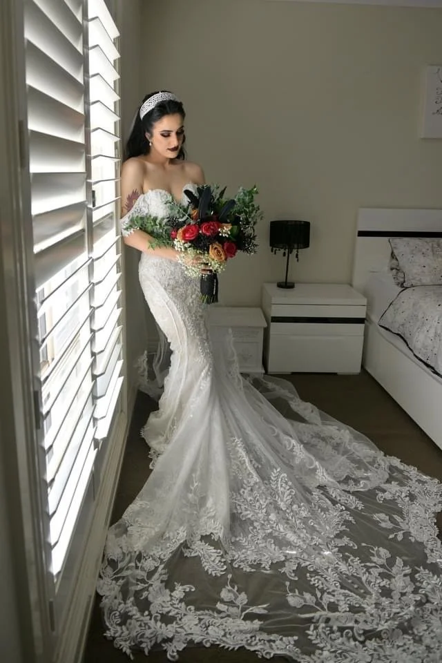 Bride in a white lace wedding dress with a long train, holding a bouquet of roses in front of a window with shutters in a bedroom.