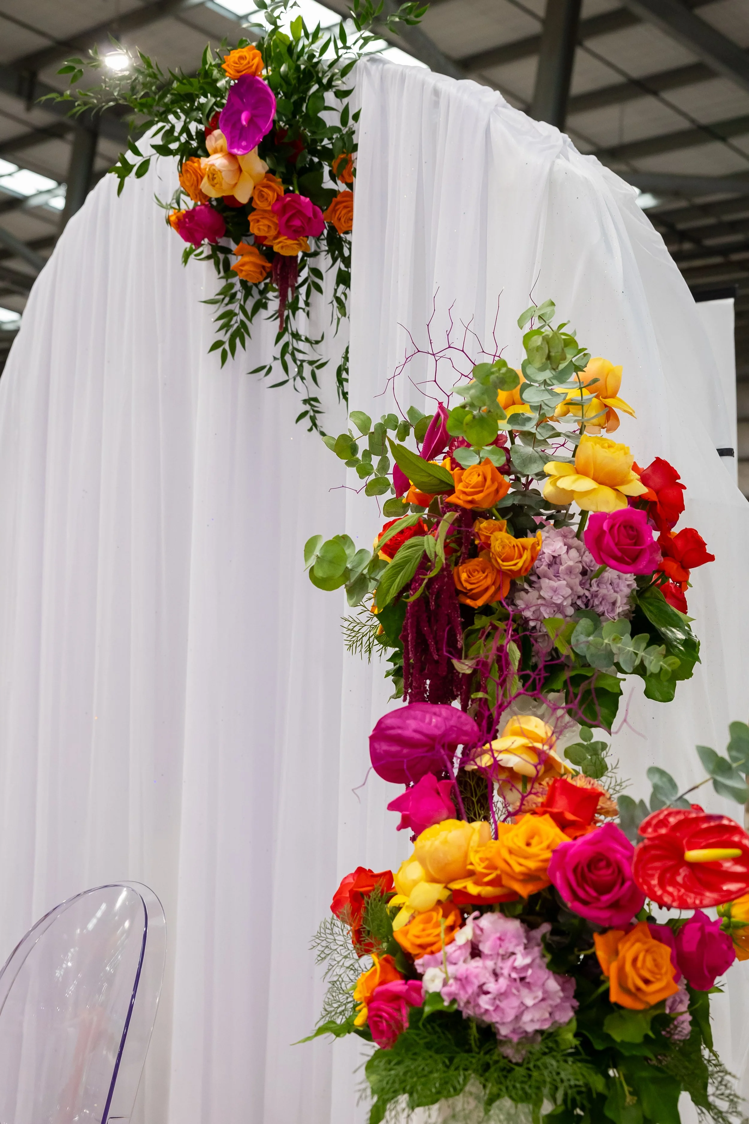 A decorative floral arrangement with colorful flowers including roses, orchids, and hydrangeas, set against a white draped curtain background, possibly for a wedding or event.