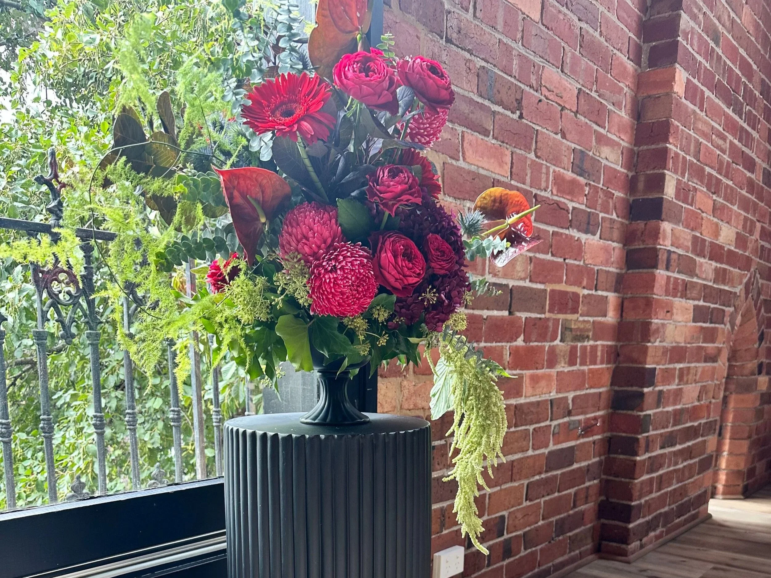 A floral arrangement of pink and red flowers in a black vase on a black pedestal near a window with a brick wall background.