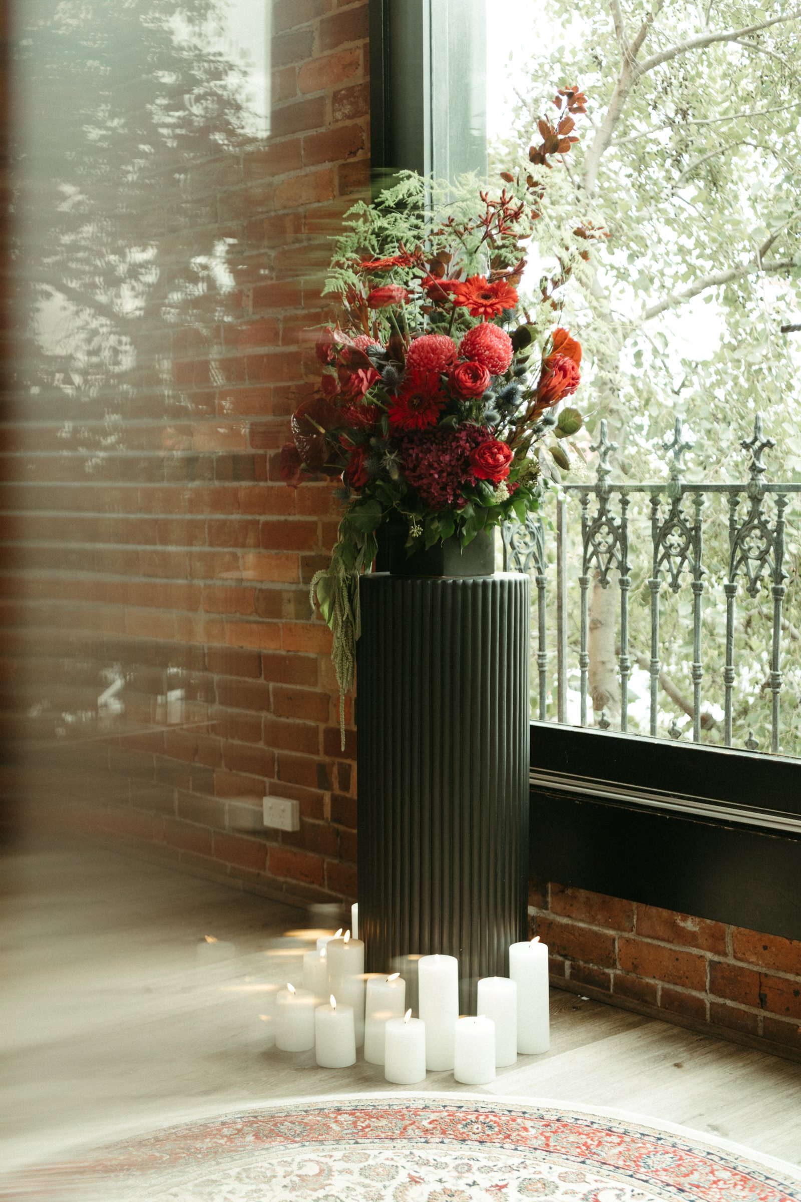 A tall black pedestal with a textured vertical stripe pattern holds a vibrant bouquet of red, pink, and orange flowers. White candles are arranged around the pedestal on the floor. Behind, there is a large window with a view of trees and an iron balcony railing outside, and a red brick wall on the inside.