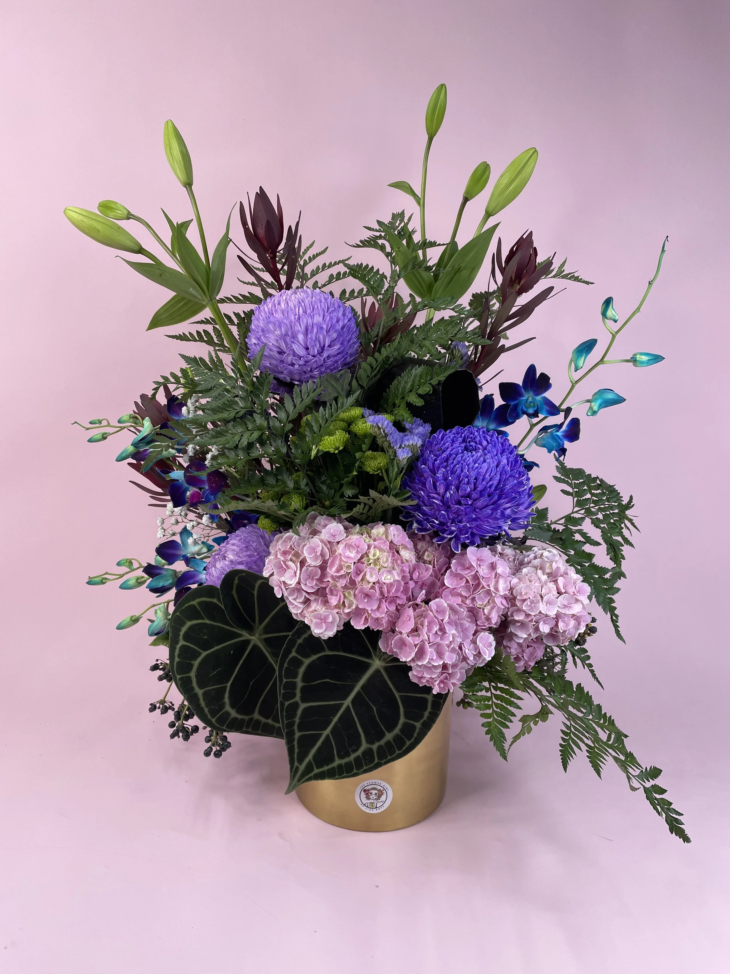 A floral arrangement in a gold pot with purple, pink, and blue flowers, green leaves, and ferns, set against a pink background.