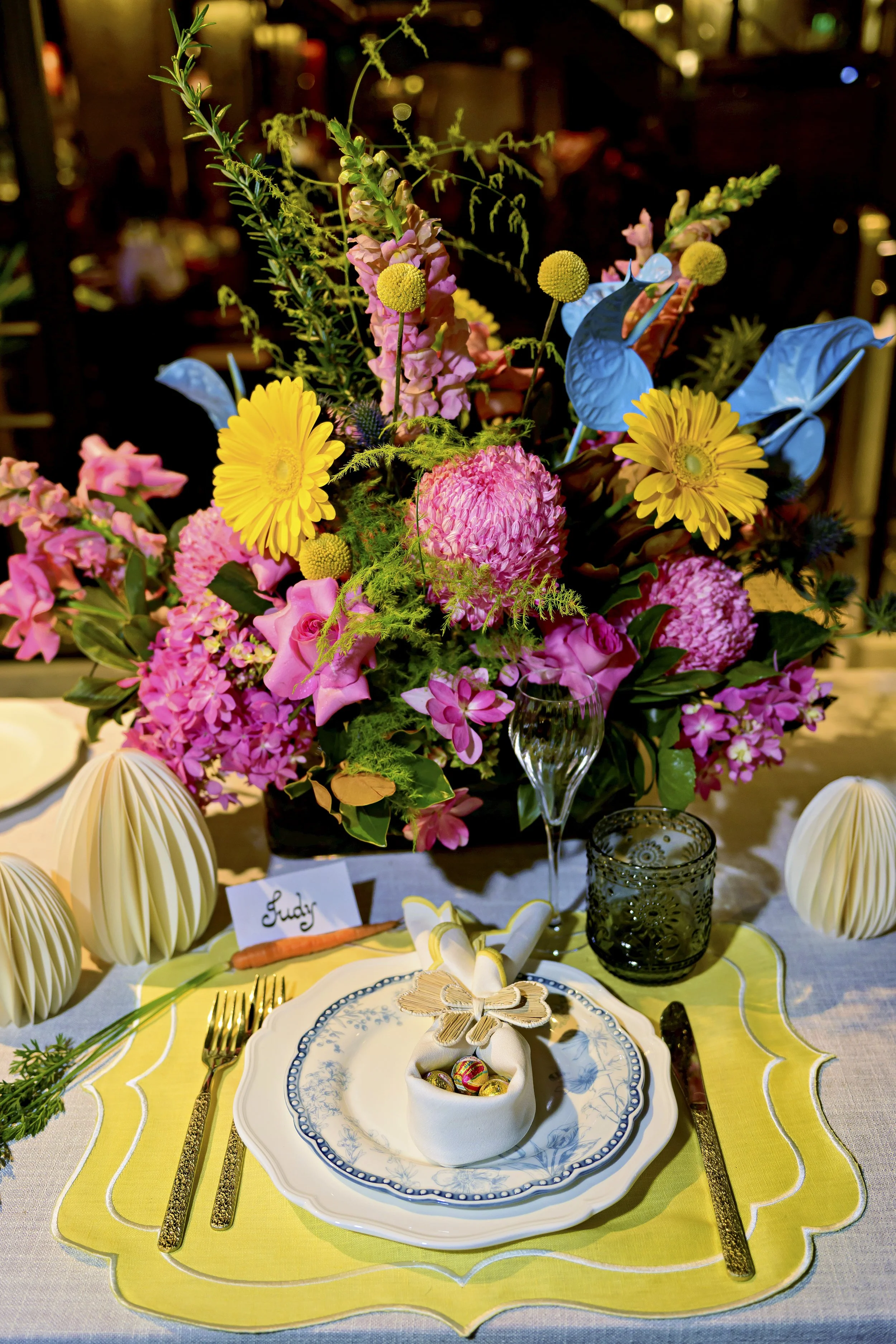 Elegant table setting with a large colorful flower arrangement, decorative white folded paper lanterns, a plate with a butterfly-shaped napkin, colorful painted eggs, gold cutlery, a wine glass, and candles.