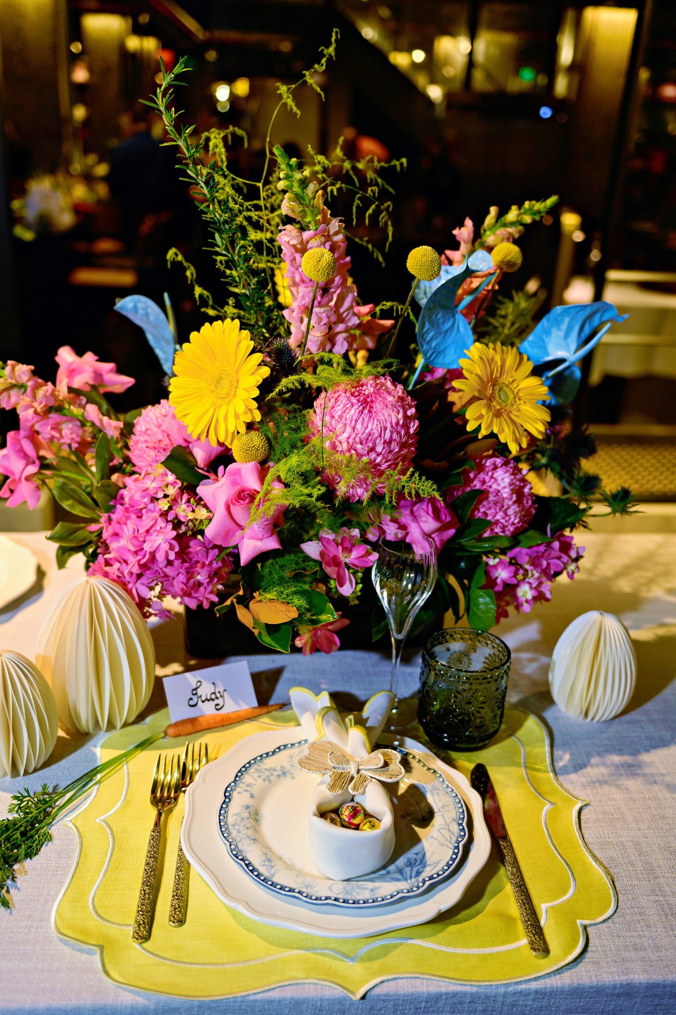 A table setting with a large colorful flower arrangement in the center, surrounded by white and yellow plates, gold utensils, and decorative paper lanterns, at a festive event.