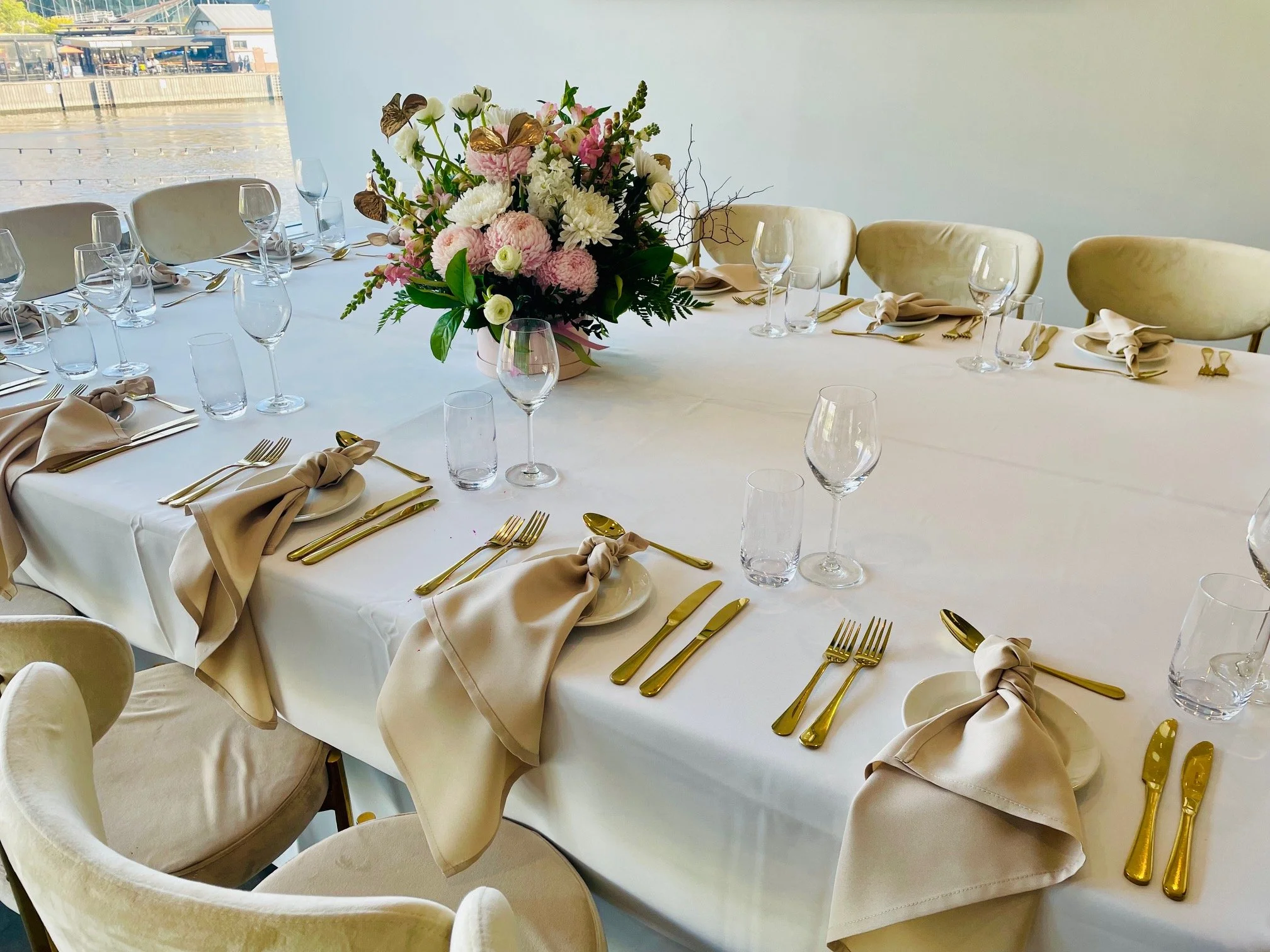 Elegant banquet table with a white tablecloth, gold utensils, beige napkins tied on plates, water glasses, wine glasses, and a floral centerpiece with pink and white flowers.