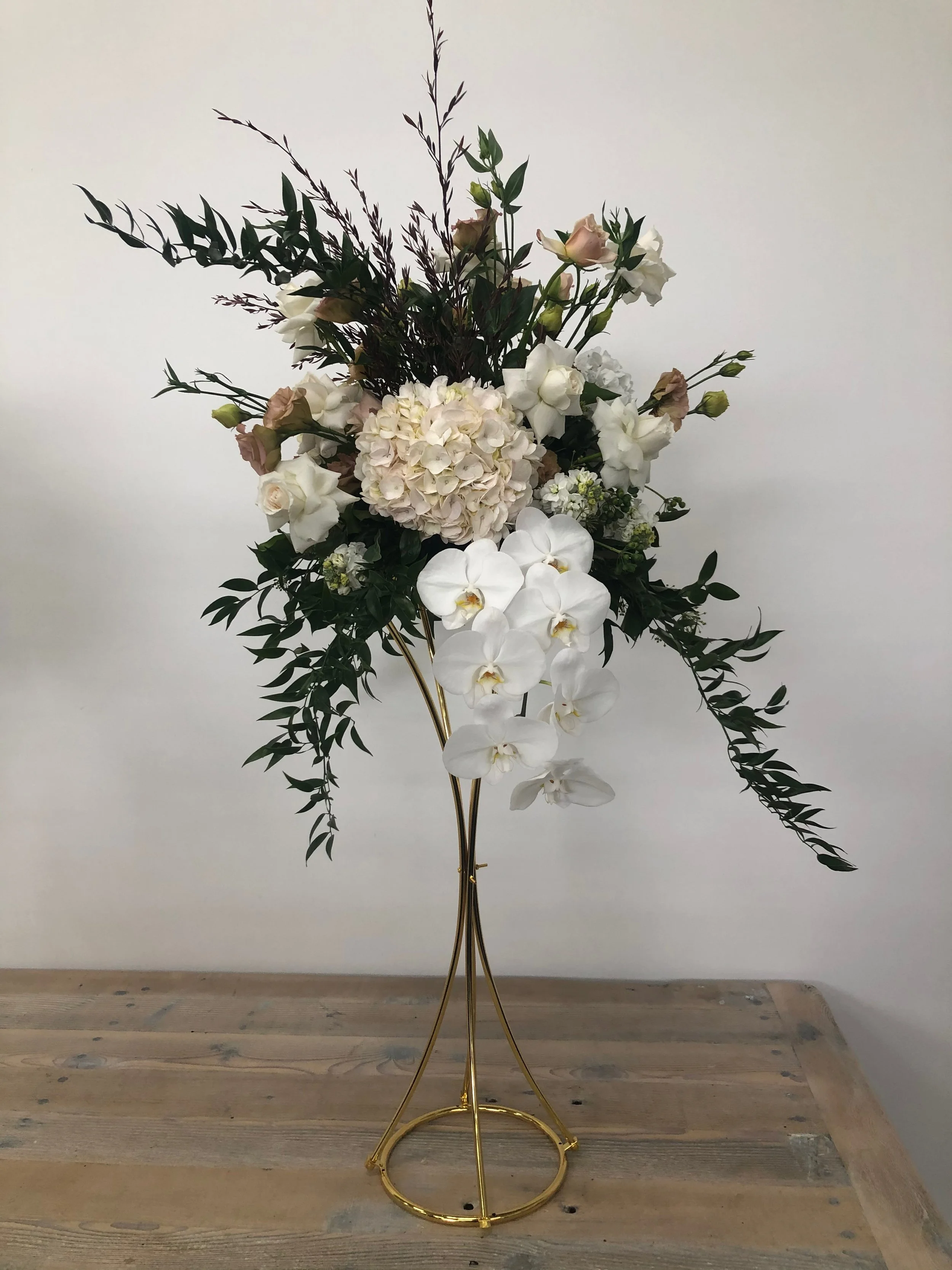 A tall floral arrangement with white orchids, pale pink roses, hydrangeas, and various greenery, displayed in a gold stand on a wooden surface.
