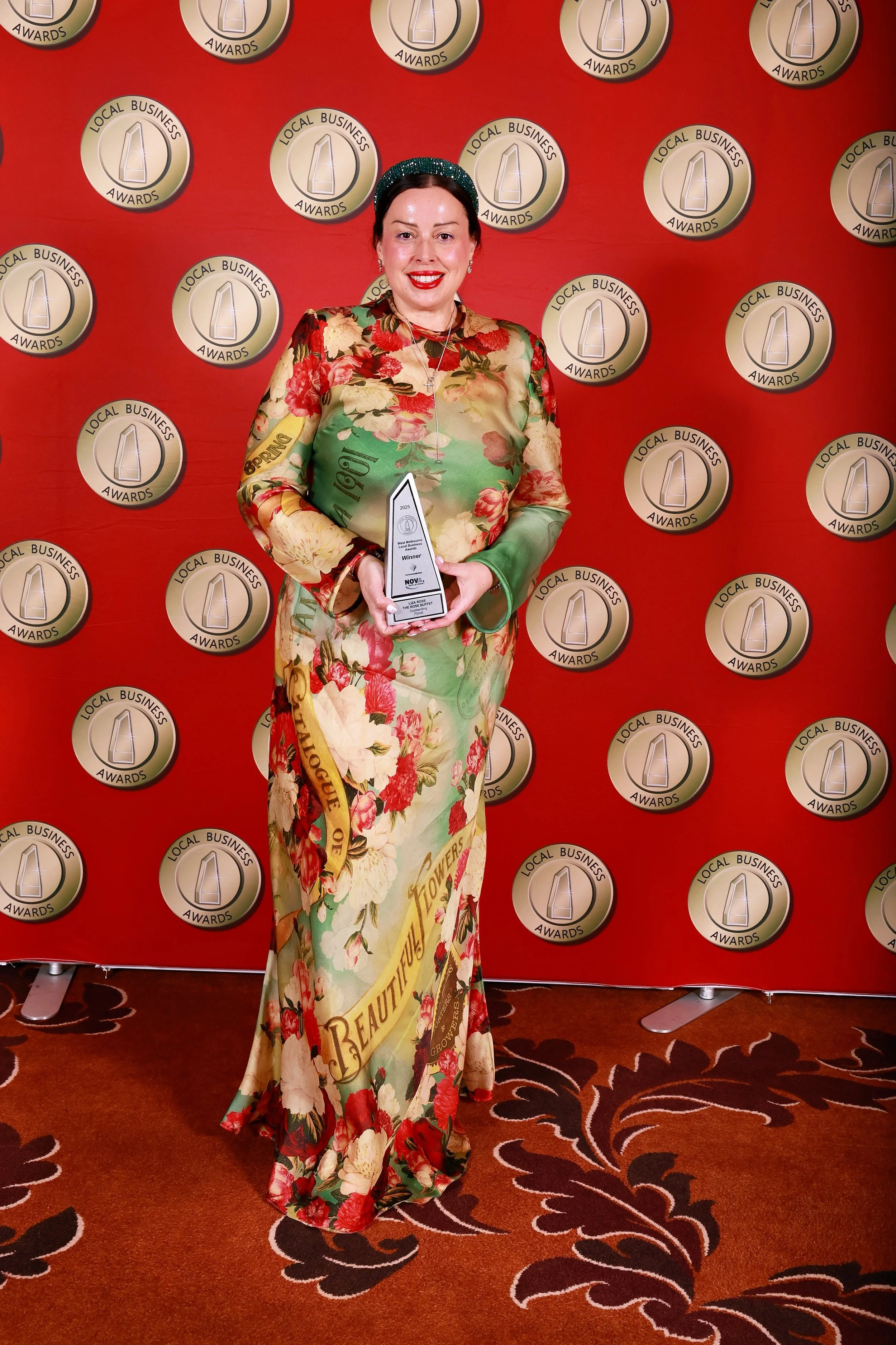 Liza Rose dressed in a floral gown holding a trophy at the Local Business Awards ceremony, standing in front of a red backdrop with the event logo.