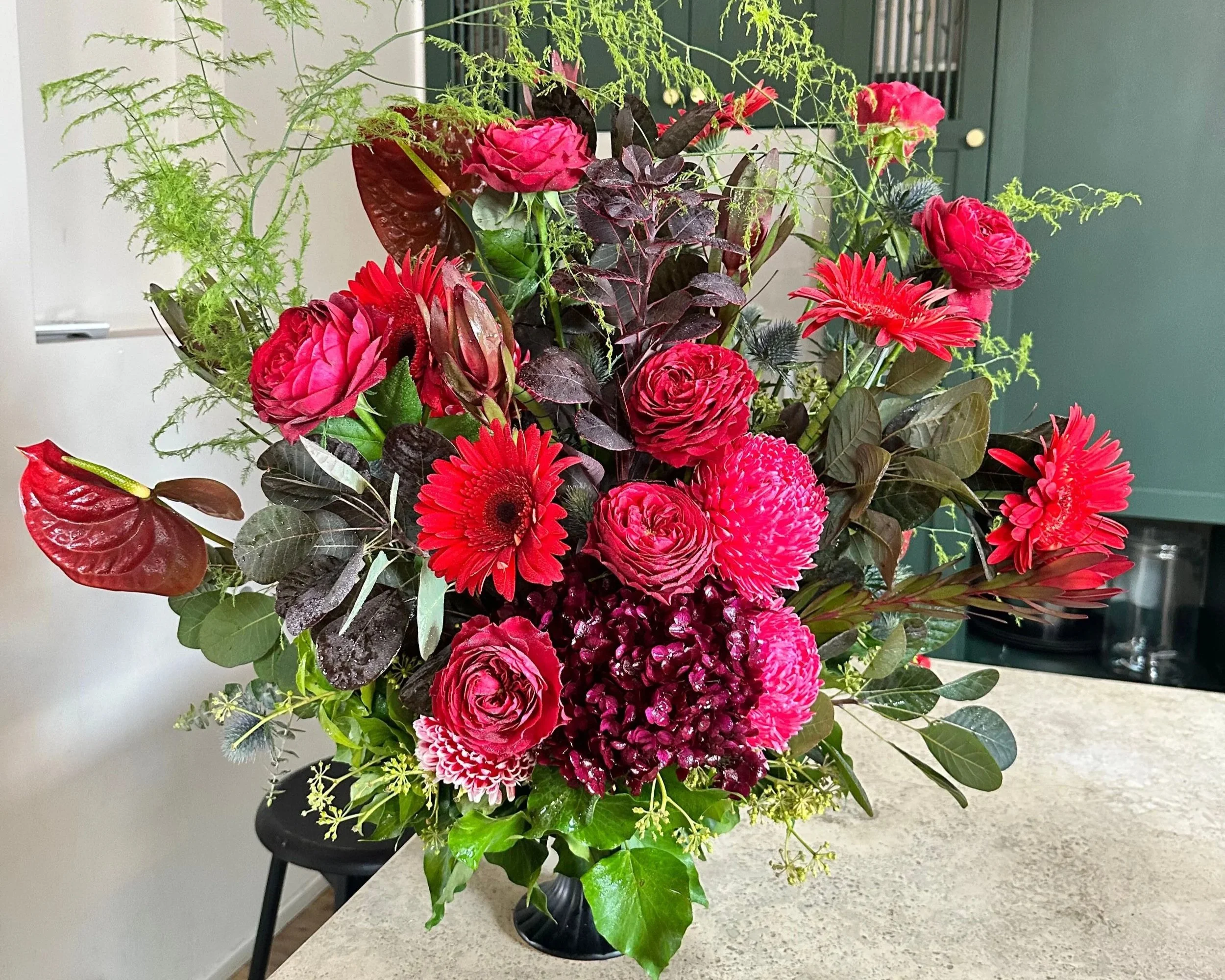 A vibrant bouquet of pink and red flowers with green and dark burgundy foliage in a black vase on a beige countertop.