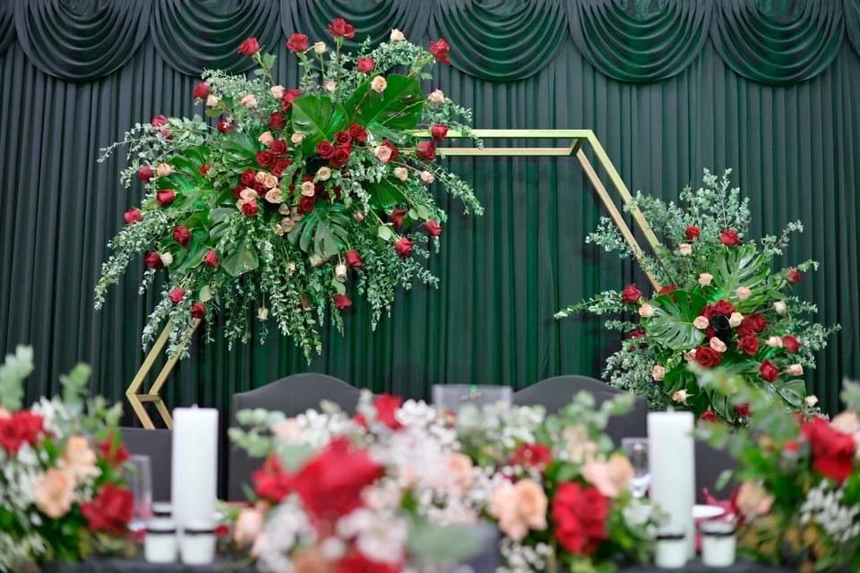 Floral arrangement with red and pink roses and greenery on a geometric gold frame in front of green curtains.