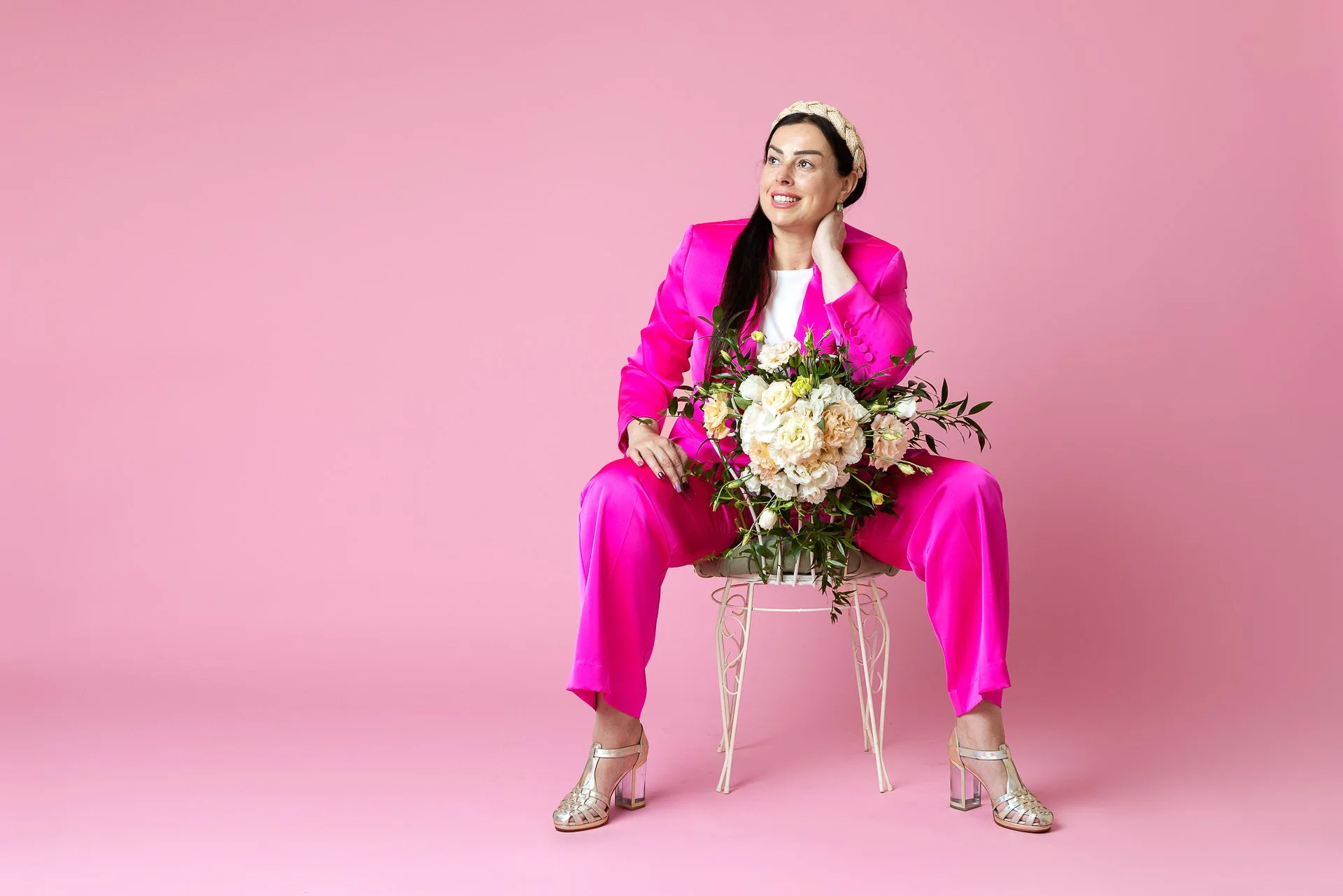 Woman sitting on a small stool, wearing a bright pink suit, holding a bouquet of white flowers, with a pink background.