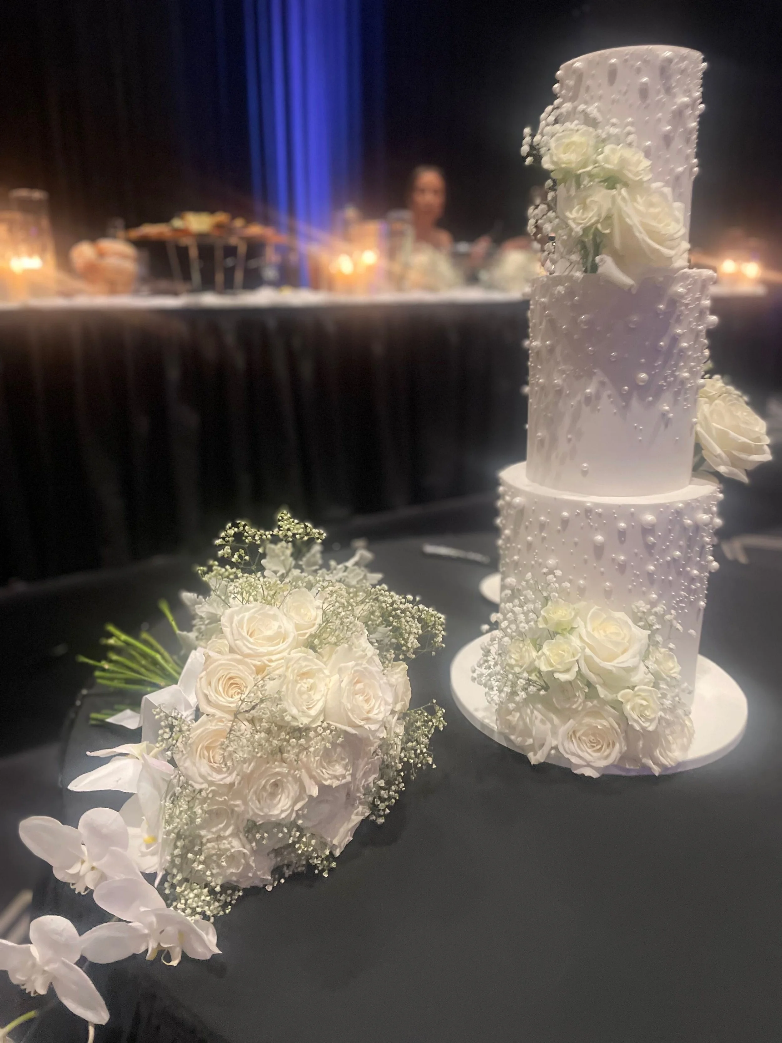 Wedding cake decorated with white flowers and pearls, placed on a black table with a matching bouquet of white roses and baby's breath.