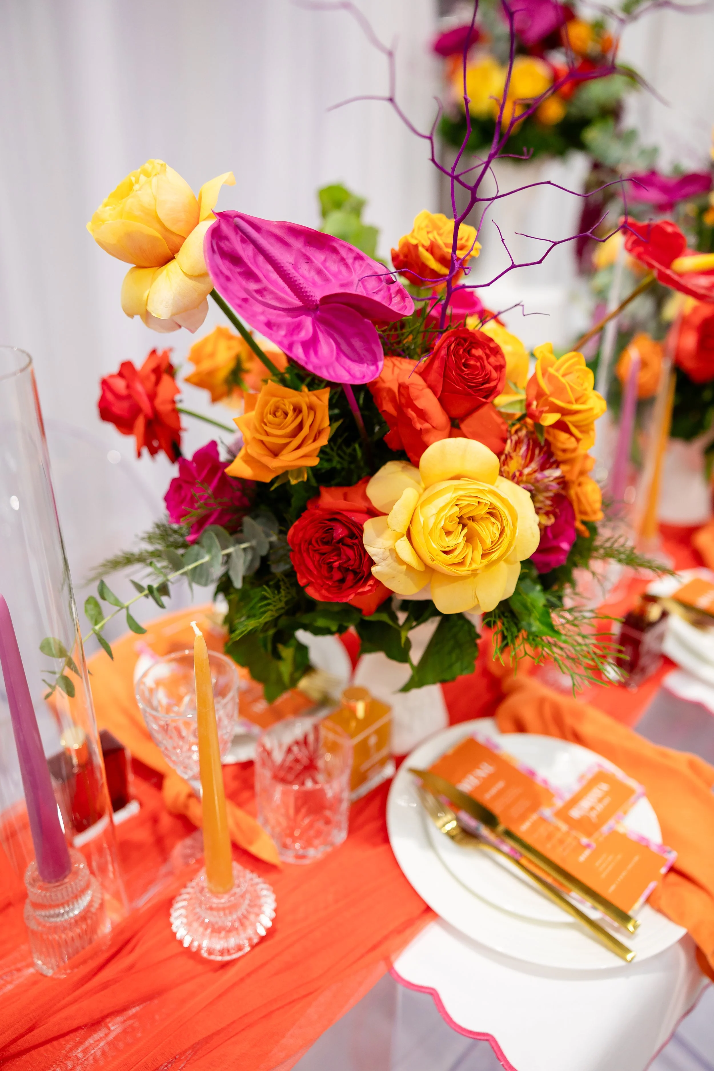 Colorful floral centerpiece with roses, anthuriums, and other blooms on a decorated table with orange table runner, candles, and place settings.