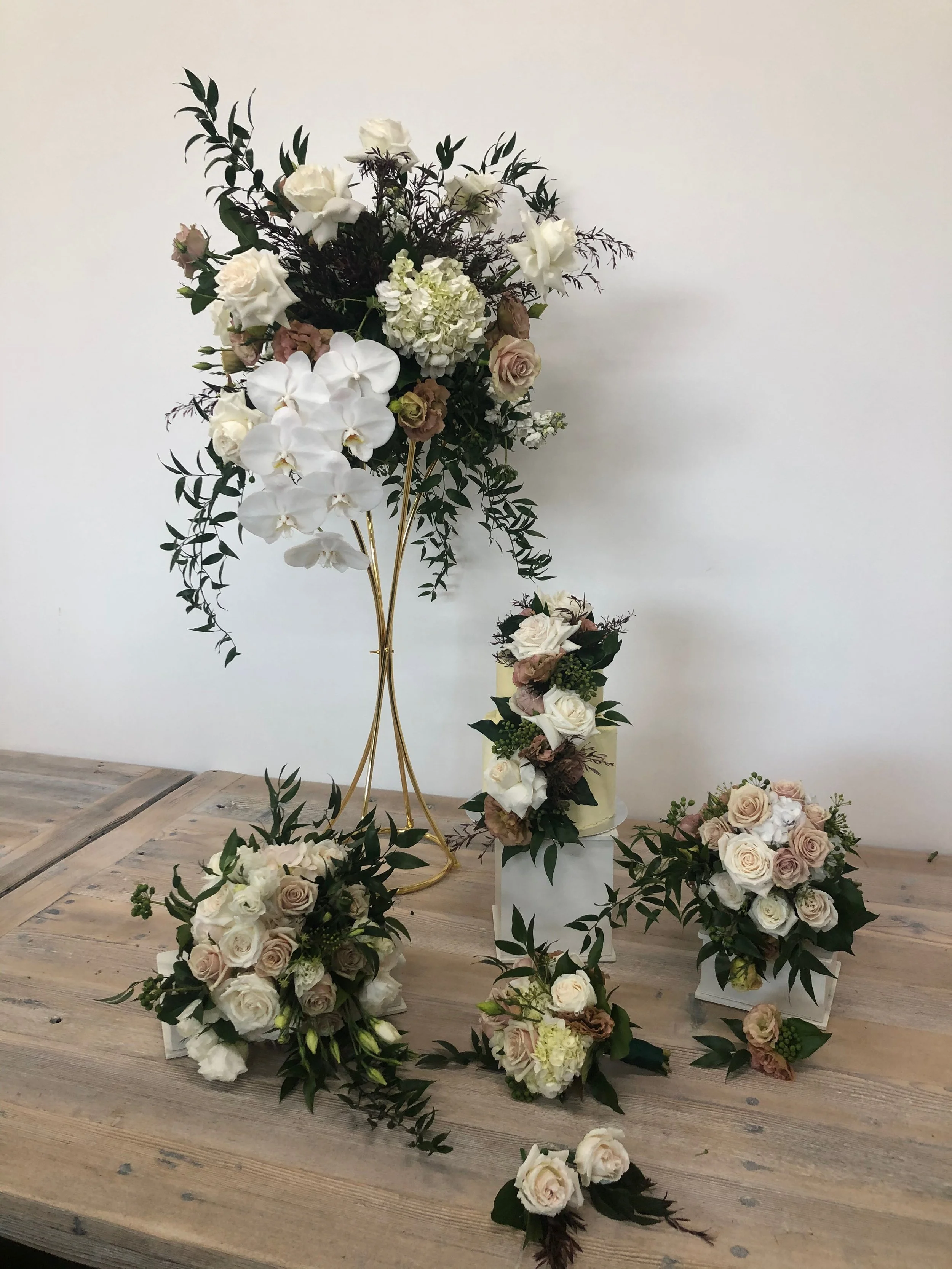 Arrangement of white and blush roses, white orchids, hydrangeas, greenery, and other flowers on a wooden table, including a tall floral display on a gold stand and several smaller bouquets and arrangements.