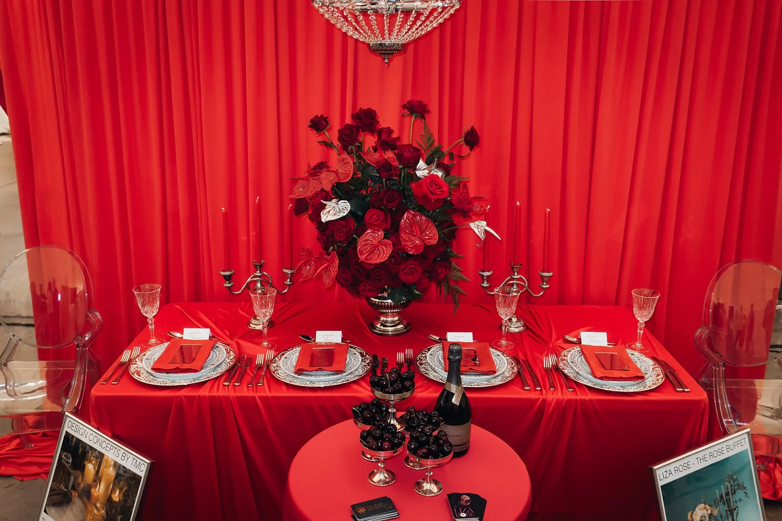 A red-themed table setting for a special occasion, with a large central floral arrangement of red roses, anthuriums, and butterflies, surrounded by silver candelabras, fine china, red napkins, and glassware, with a red curtain backdrop.