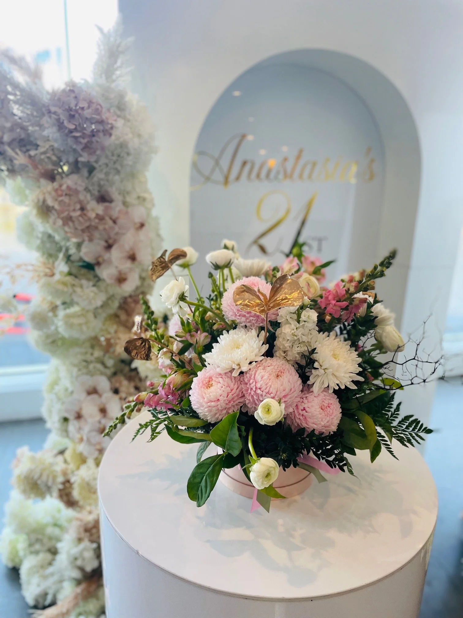 A pink floral arrangement with pink and white flowers and green leaves, set on a white cylindrical pedestal in front of a blurred background featuring a white wall with gold lettering that reads "Anastasia's 21st".