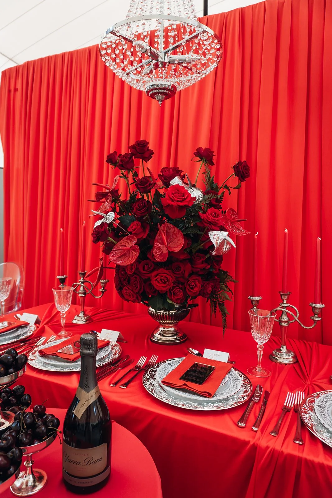 Elegant dining table with a large centerpiece of red roses and white flowers, set against red drapes. The table features silver and glass dinnerware, silver candelabras, wine glasses, a bottle of sparkling wine, and red napkins, creating a luxurious, romantic atmosphere.