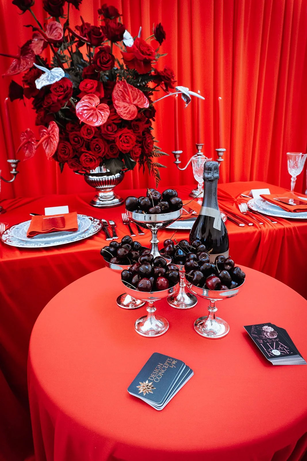 Table decorated with red tablecloth, a large flower arrangement of red roses and anthuriums, a bottle of champagne, multiple bowls of cherries, and elegant glassware. The backdrop is red curtains.