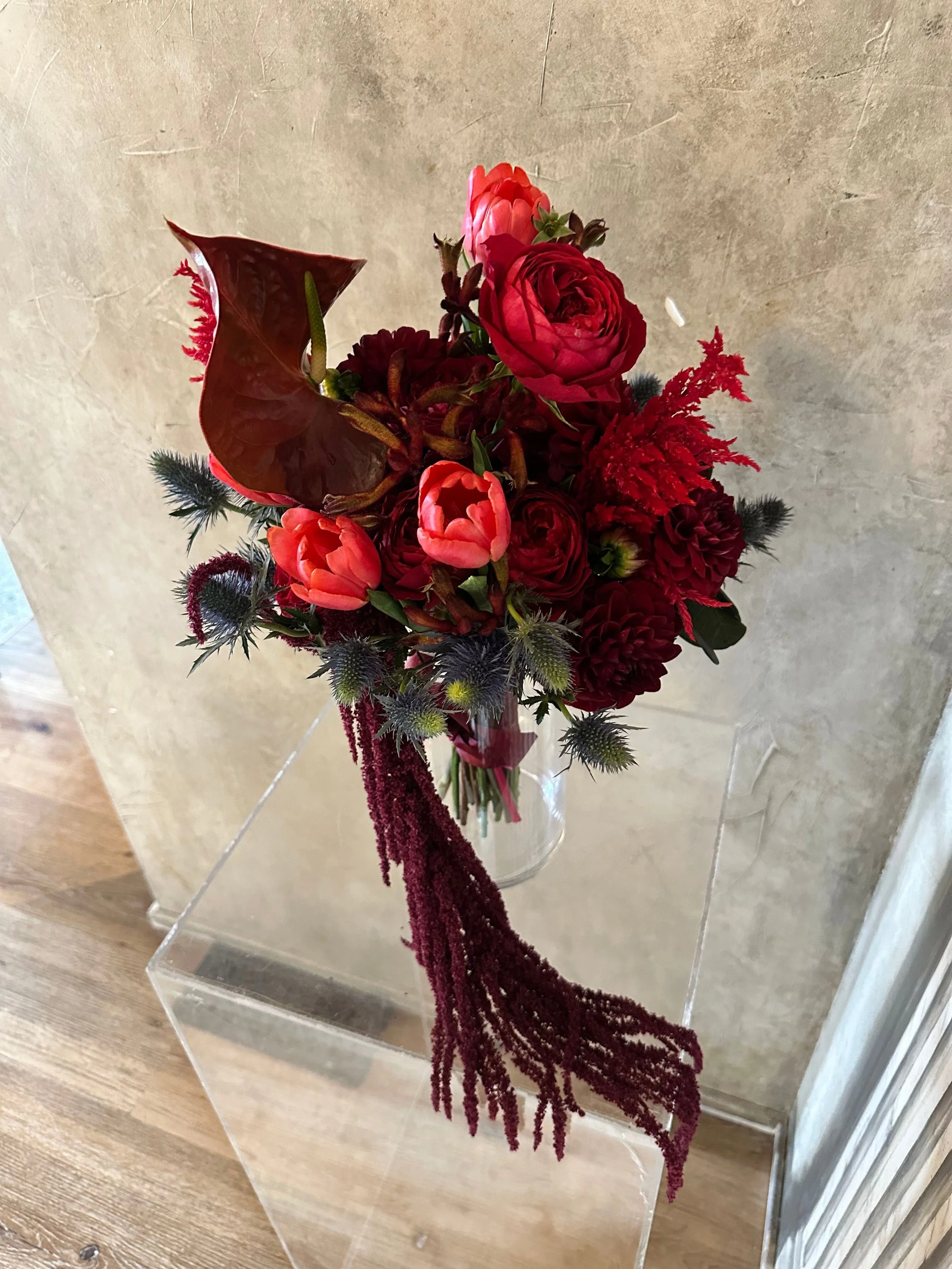 A floral arrangement with red and pink flowers, dark purple foliage, and cascading dark red amaranthus in a clear glass vase on a transparent stand.