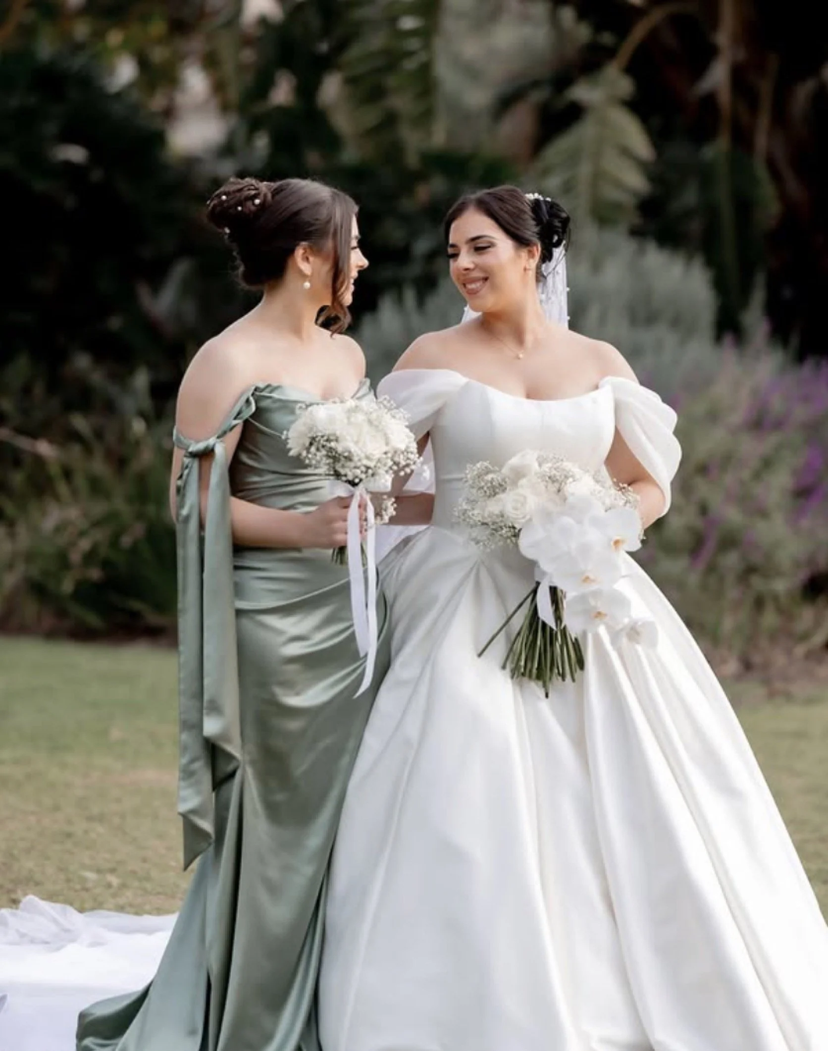 Two women in wedding attire, one in a white wedding dress and the other in a green bridesmaid dress, holding bouquets, standing on a grassy area outdoor with trees in the background.