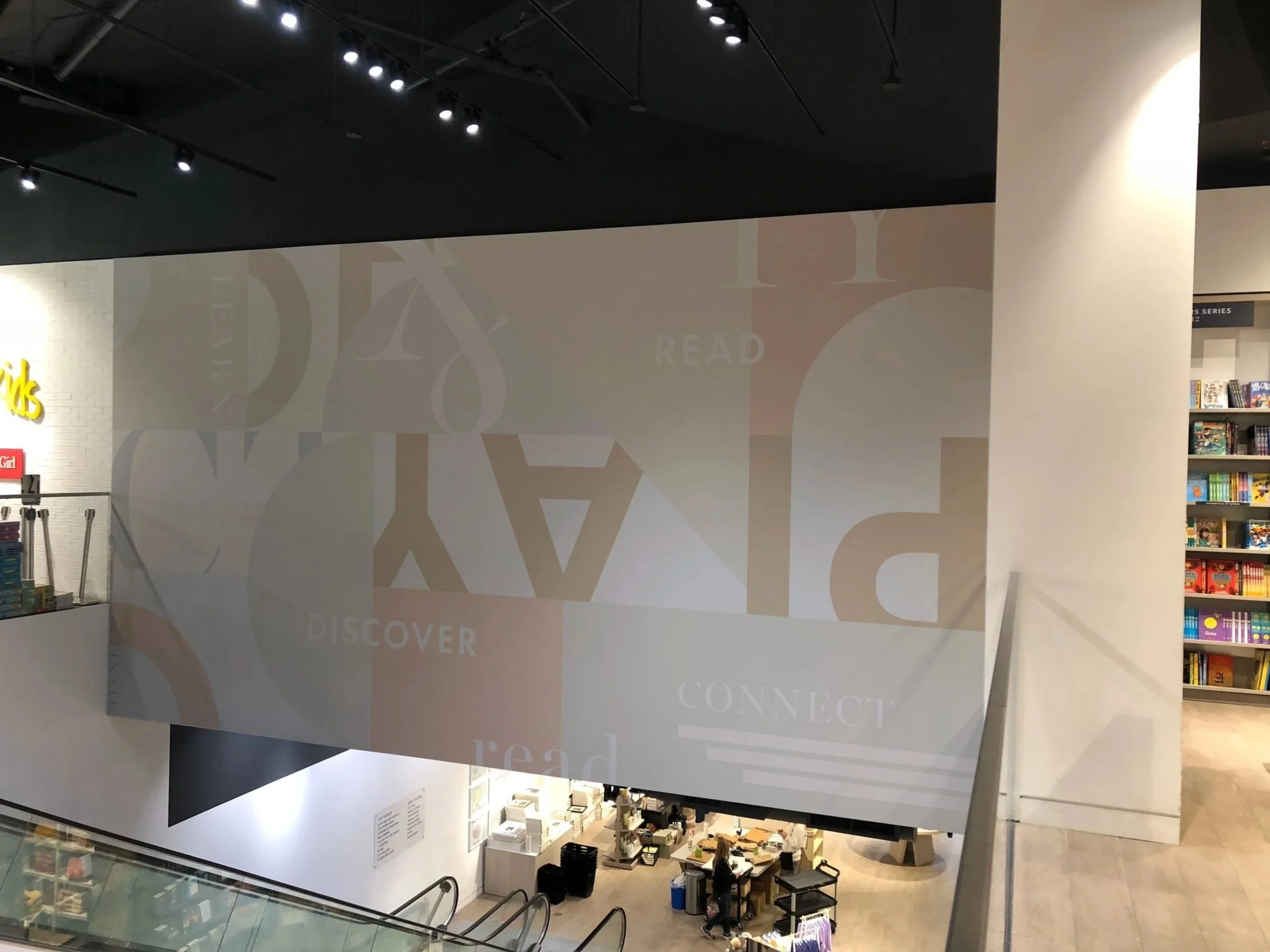 Interior of a bookstore or library with a large wall display featuring words like "READ," "DISCOVER," and "CONNECT" in various fonts and colors.