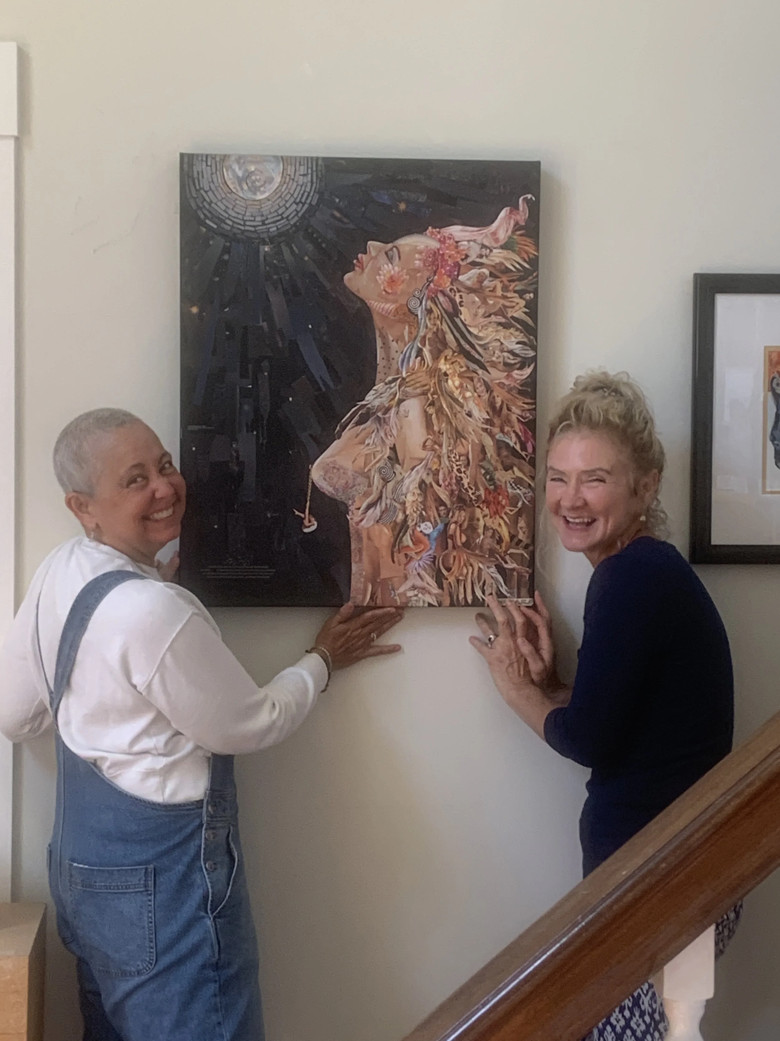 Two women are smiling and hanging a colorful artwork of a woman with flowing hair and feathers on a white wall.