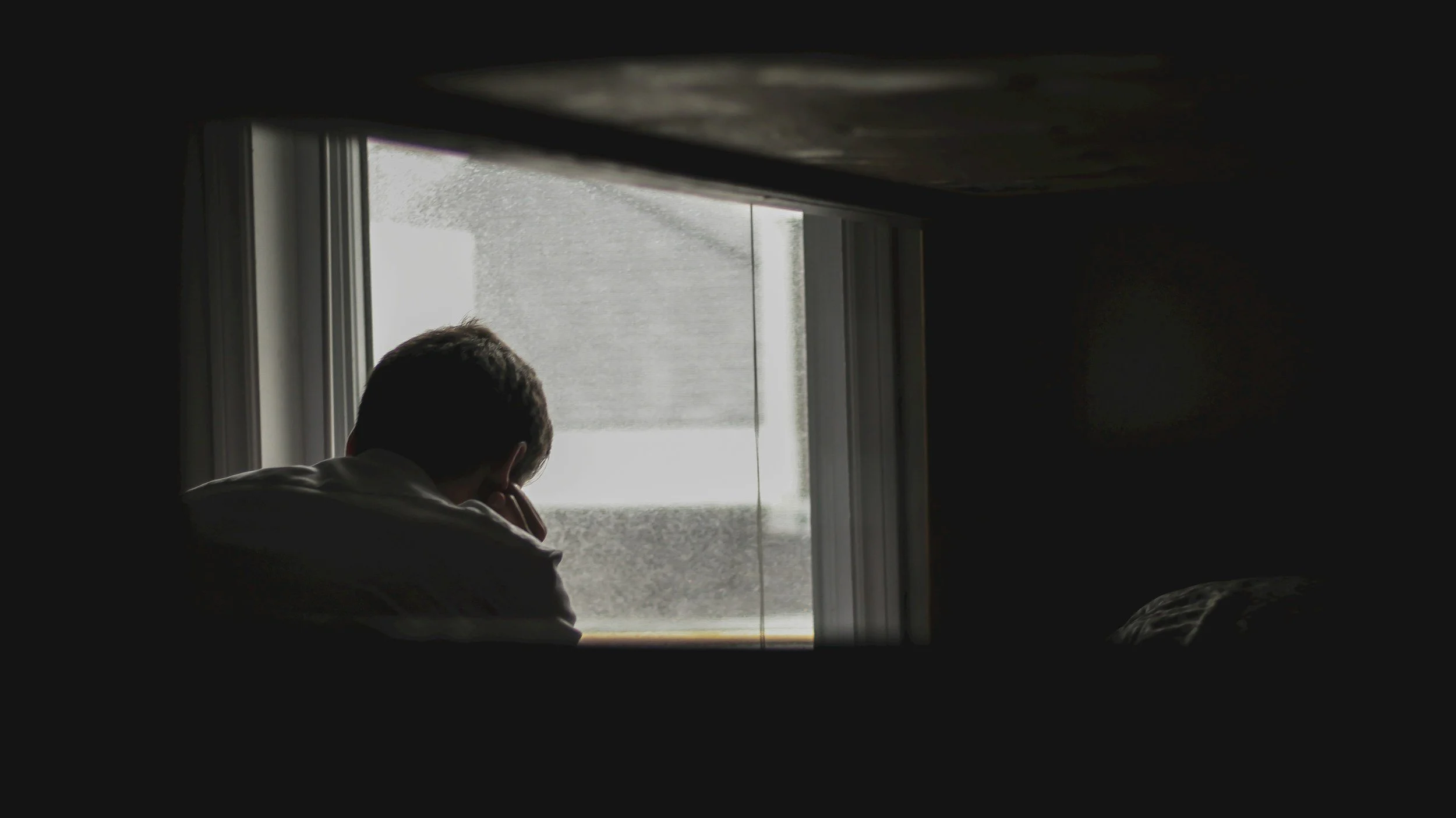 A person sitting by a window, resting their head on their hand, with a view of a cloudy or foggy outdoor scene.