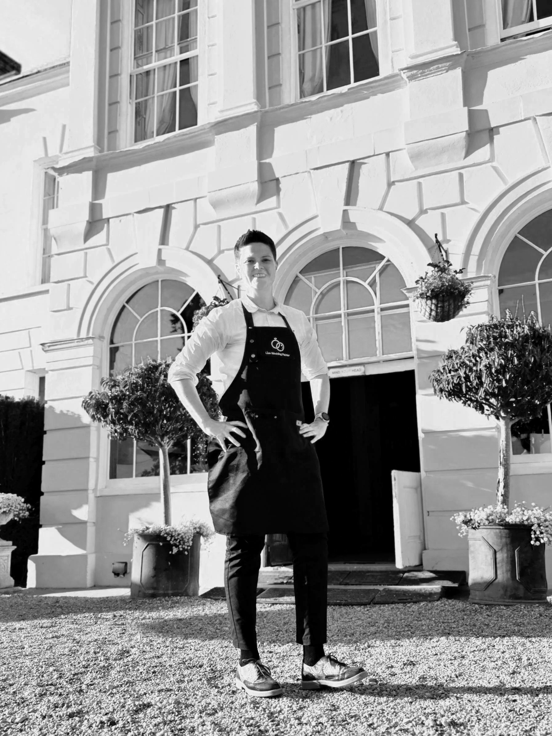 A person in casual attire wearing an apron standing outside a large historic building with arched windows and potted plants.