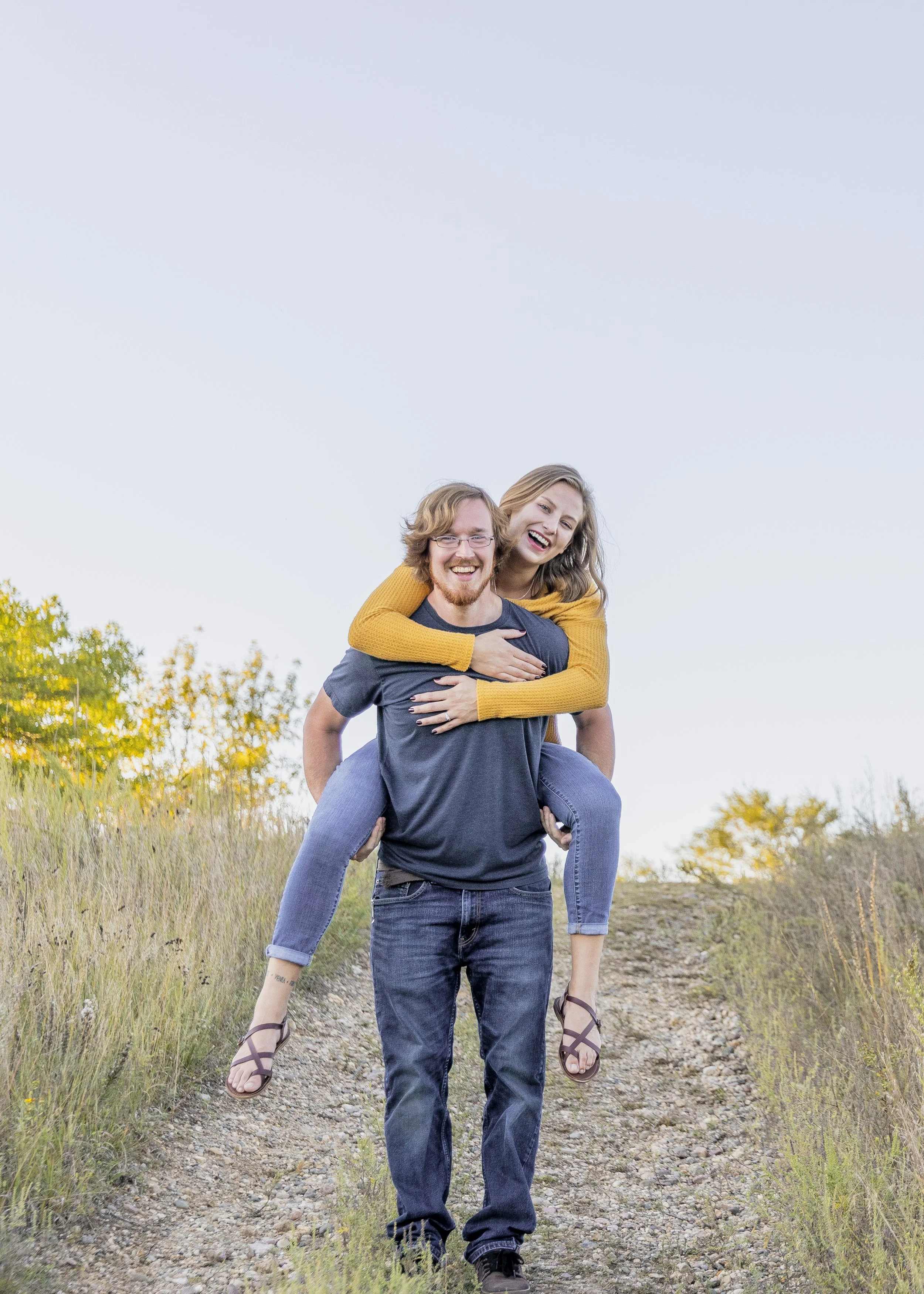 A couple outdoors on a dirt trail, with the woman on the man's back. They are smiling and laughing during the daytime.