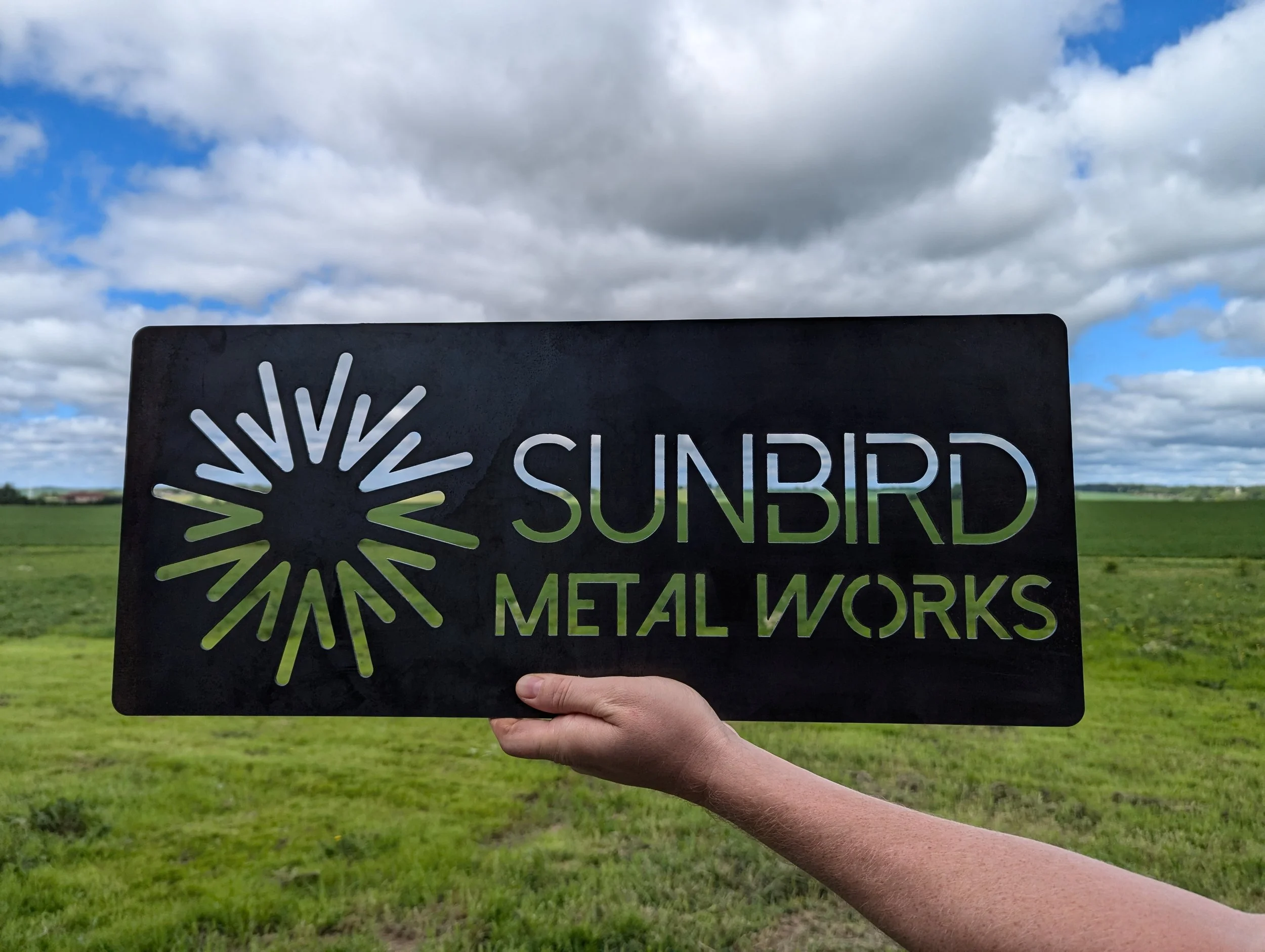 A person holding a black metal sign that reads "SUNBIRD METAL WORKS" with a sunburst logo, against a background of green fields and a partly cloudy sky.