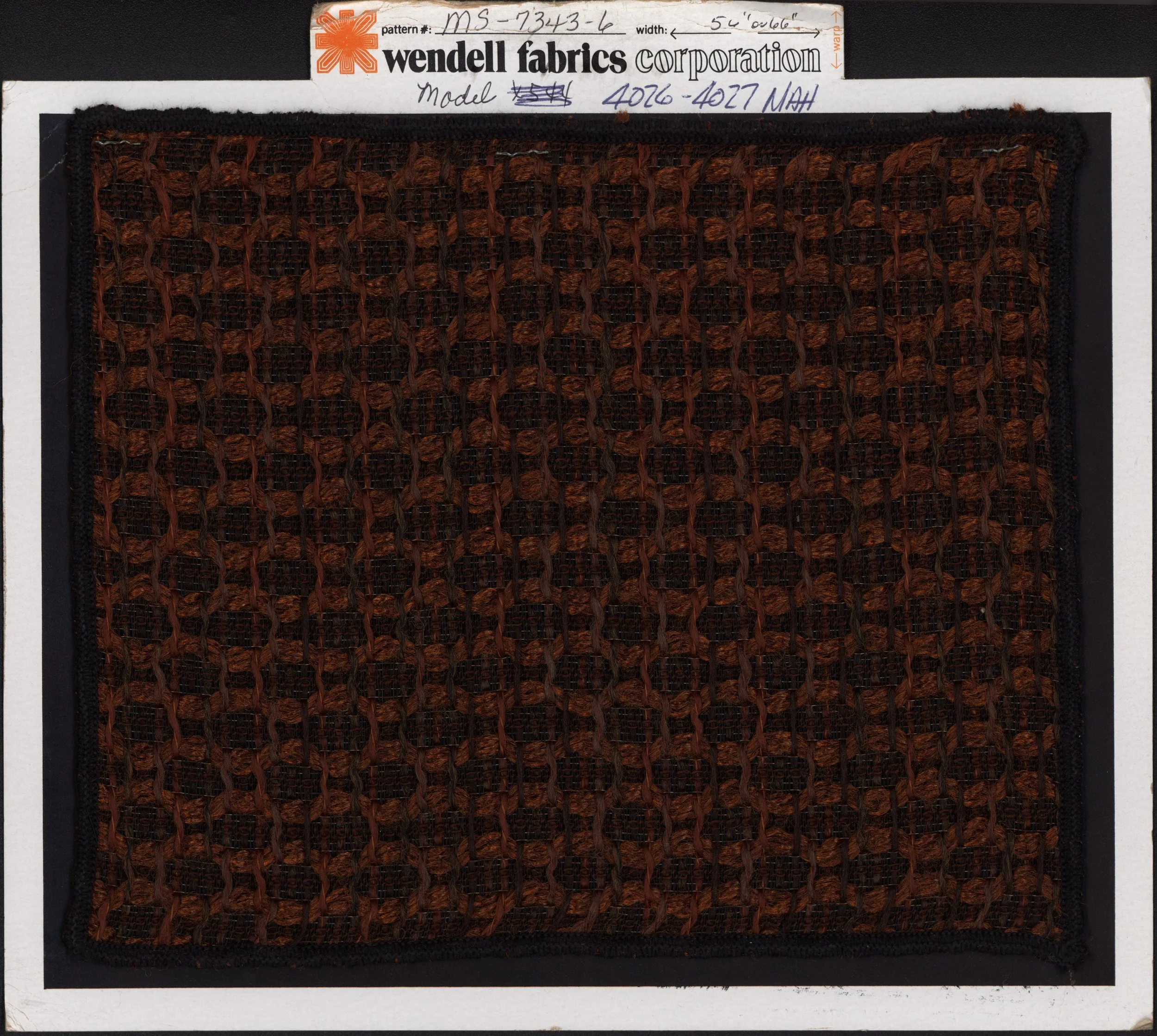 Close-up of a fabric sample from Wendell Fabrics Corporation, with a woven pattern in shades of brown and black, attached to a black backing. The sample lies beneath a white index card containing handwritten and printed information, including pattern