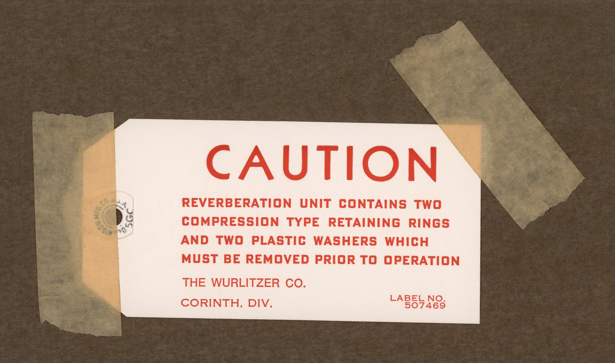 Close-up of a warning label taped to a surface, indicating caution about a reverberation unit containing compression cylinders and plastic washers that need removal before operation.