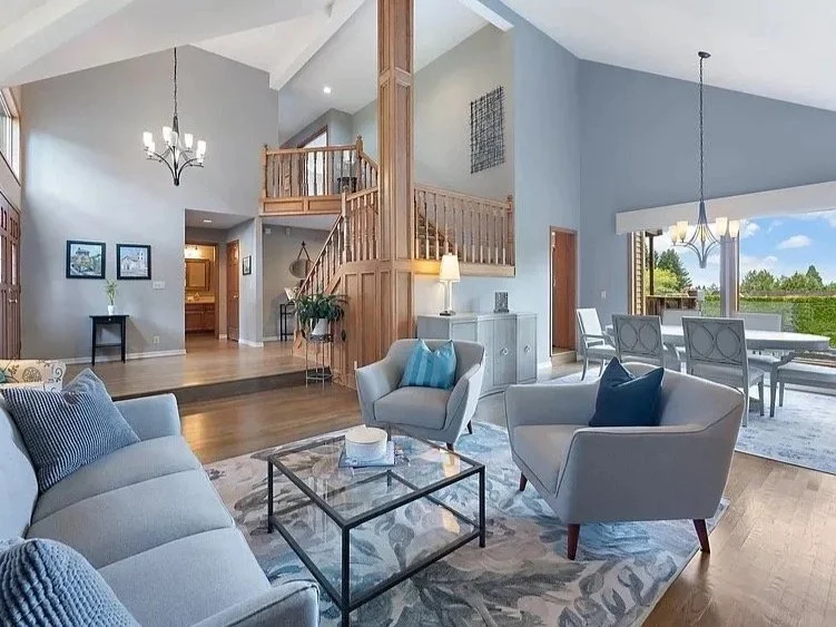 A spacious living room with light blue walls, wooden flooring, and large windows. It features two white armchairs and a sofa with blue pillows, a glass coffee table, and a patterned area rug. There is a dining area with a table and chairs near the wi