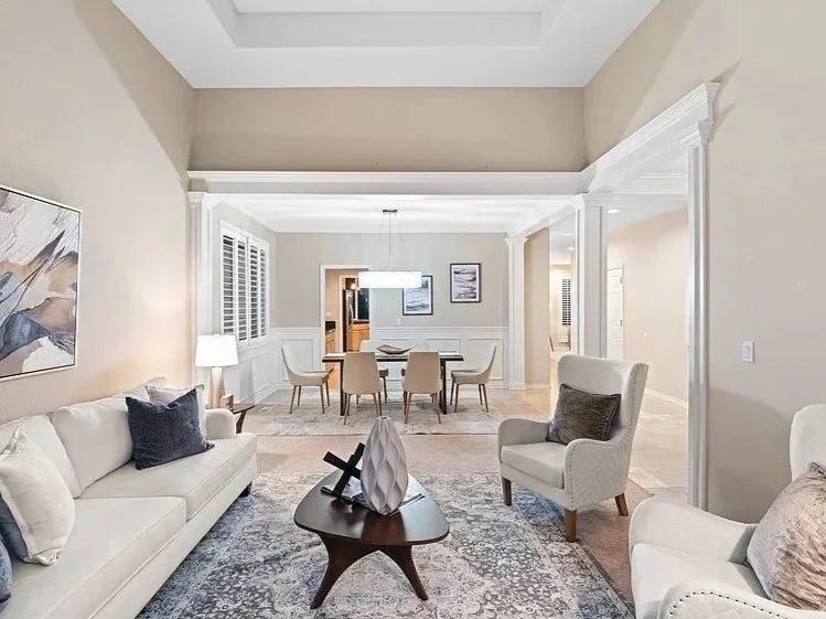 Living room and dining area with beige walls, white trim, and modern furniture. The living room has a cream sofa with throw pillows, a wooden coffee table with decorative items, and an armchair. The dining area features a rectangular table with six c