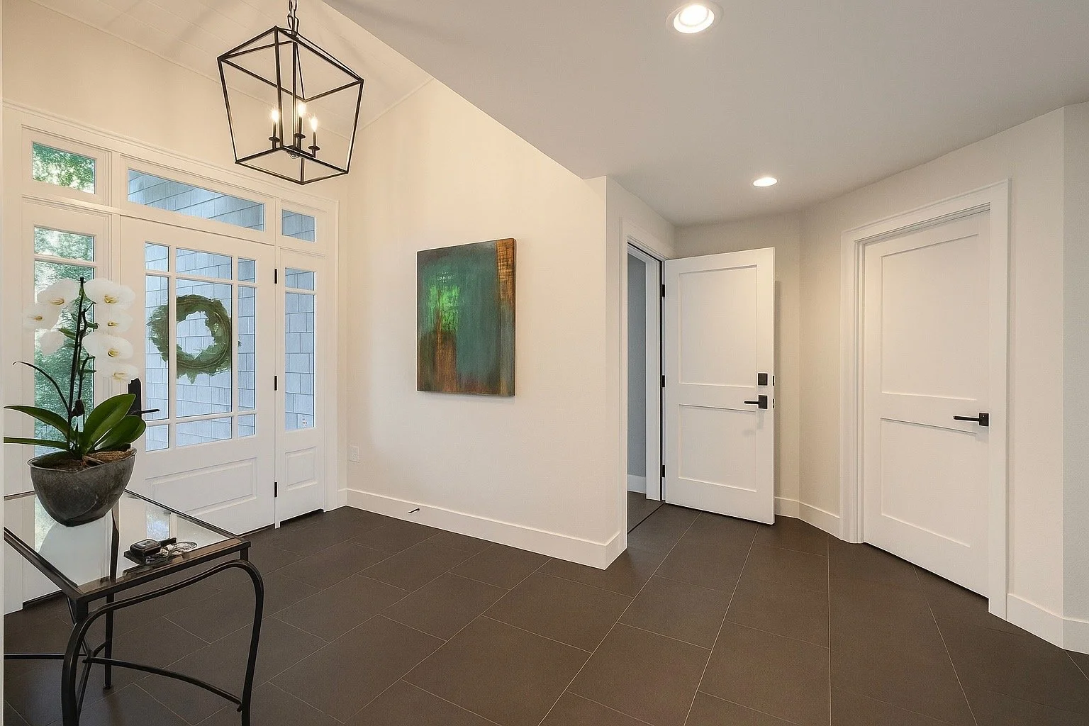 Modern minimalist interior entryway with white walls, black tile flooring, a glass and metal plant table with an orchid, a framed abstract art piece, and white double doors with glass panes.