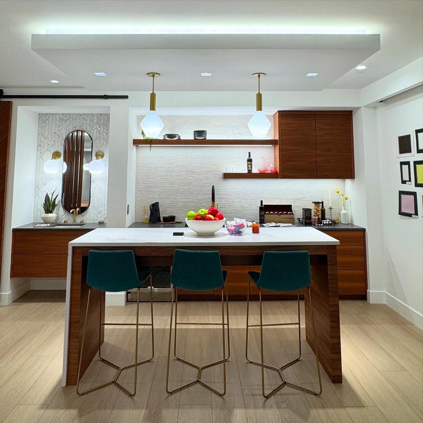 Modern kitchen with white island counter, three teal bar stools, wooden cabinetry, and wall-mounted shelves with decor. Pendant lights above the island, decorative artwork on the right wall, and a small sink area with a mirror and potted plant.