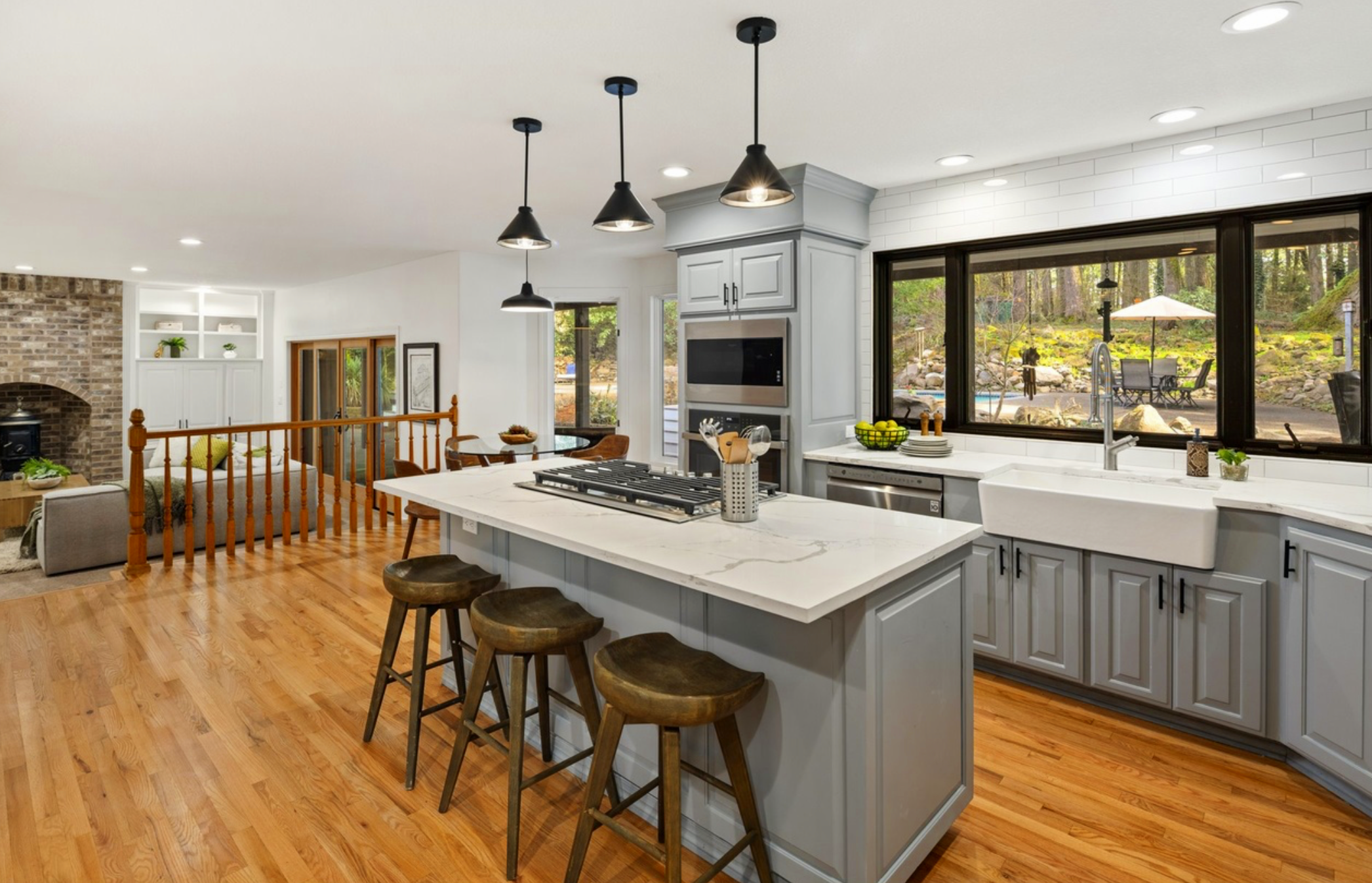 Modern kitchen with light gray cabinets, white marble countertops, a large farmhouse sink, and black pendant lights hanging over an island with three wooden stools. Large window overlooking a backyard with outdoor furniture and umbrella.