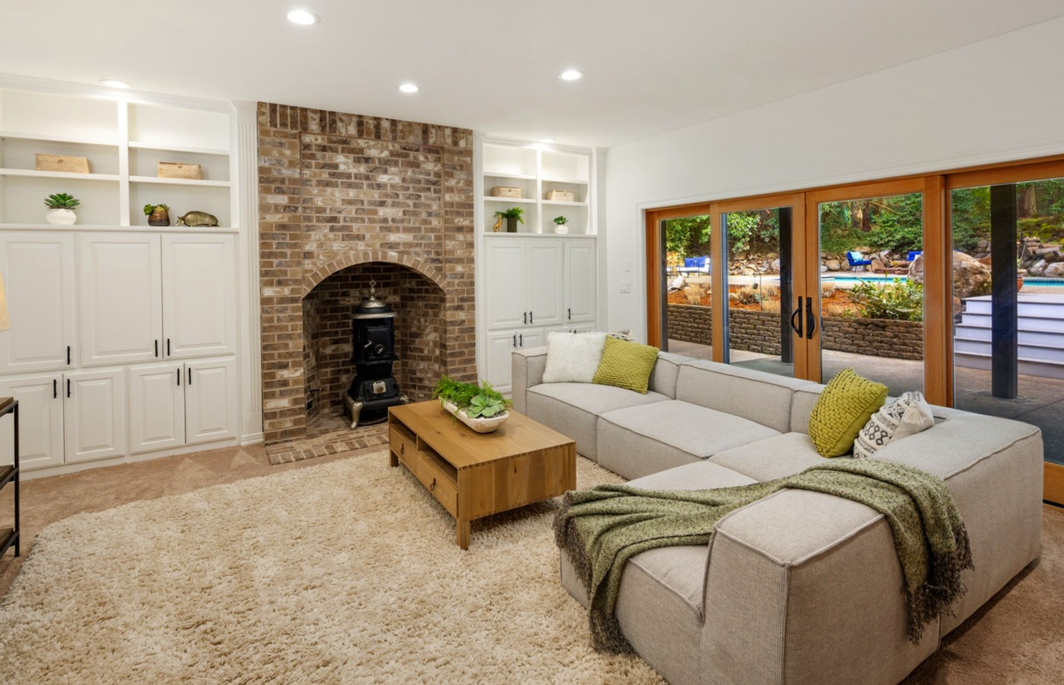 Living room with a beige sectional sofa, green and white pillows, a wooden coffee table with a plant, white built-in cabinets, brick fireplace, and large sliding glass doors leading outside.