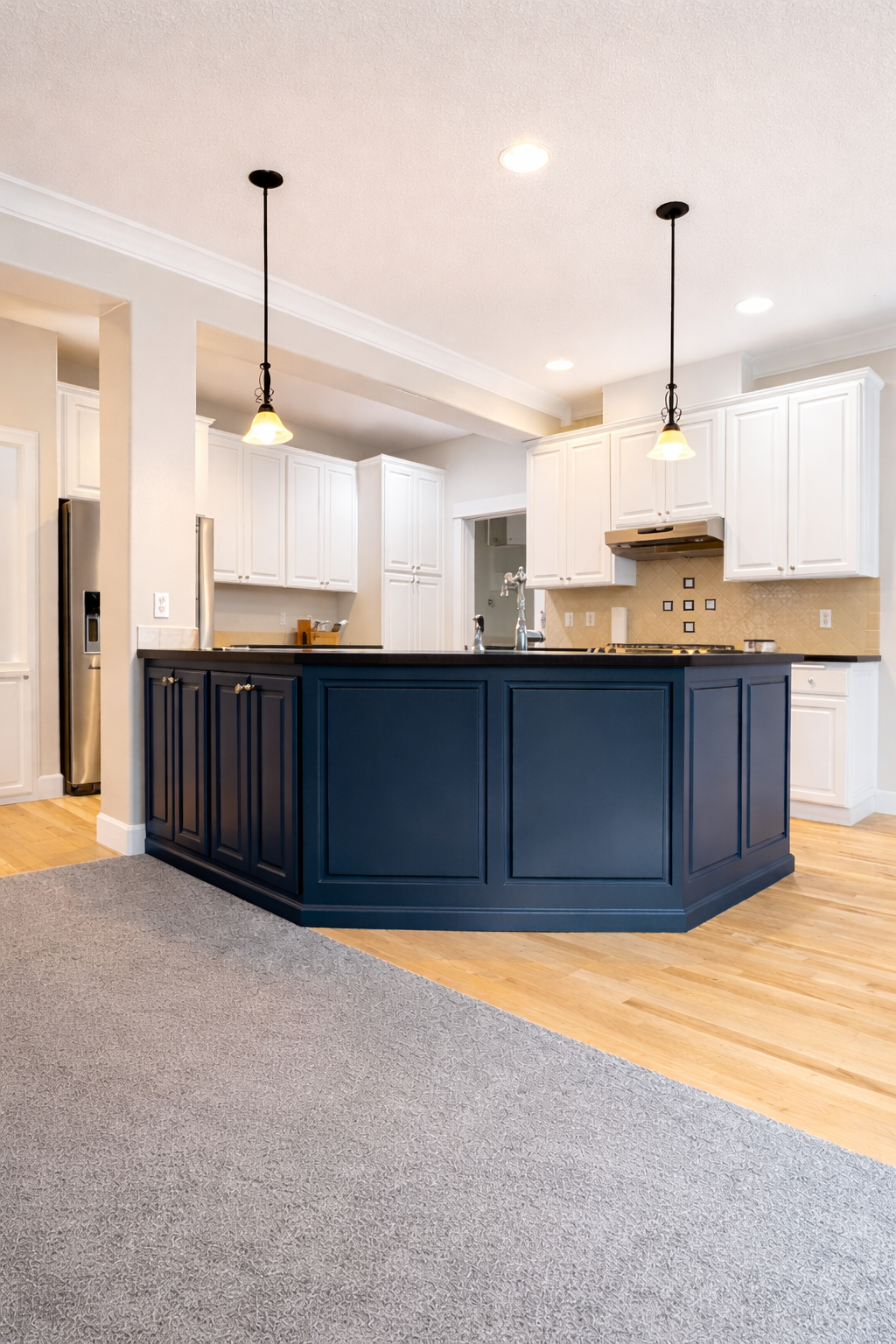 Modern kitchen with white cabinets, a navy blue island, pendant lighting, hardwood flooring, and a beige tiled backsplash.