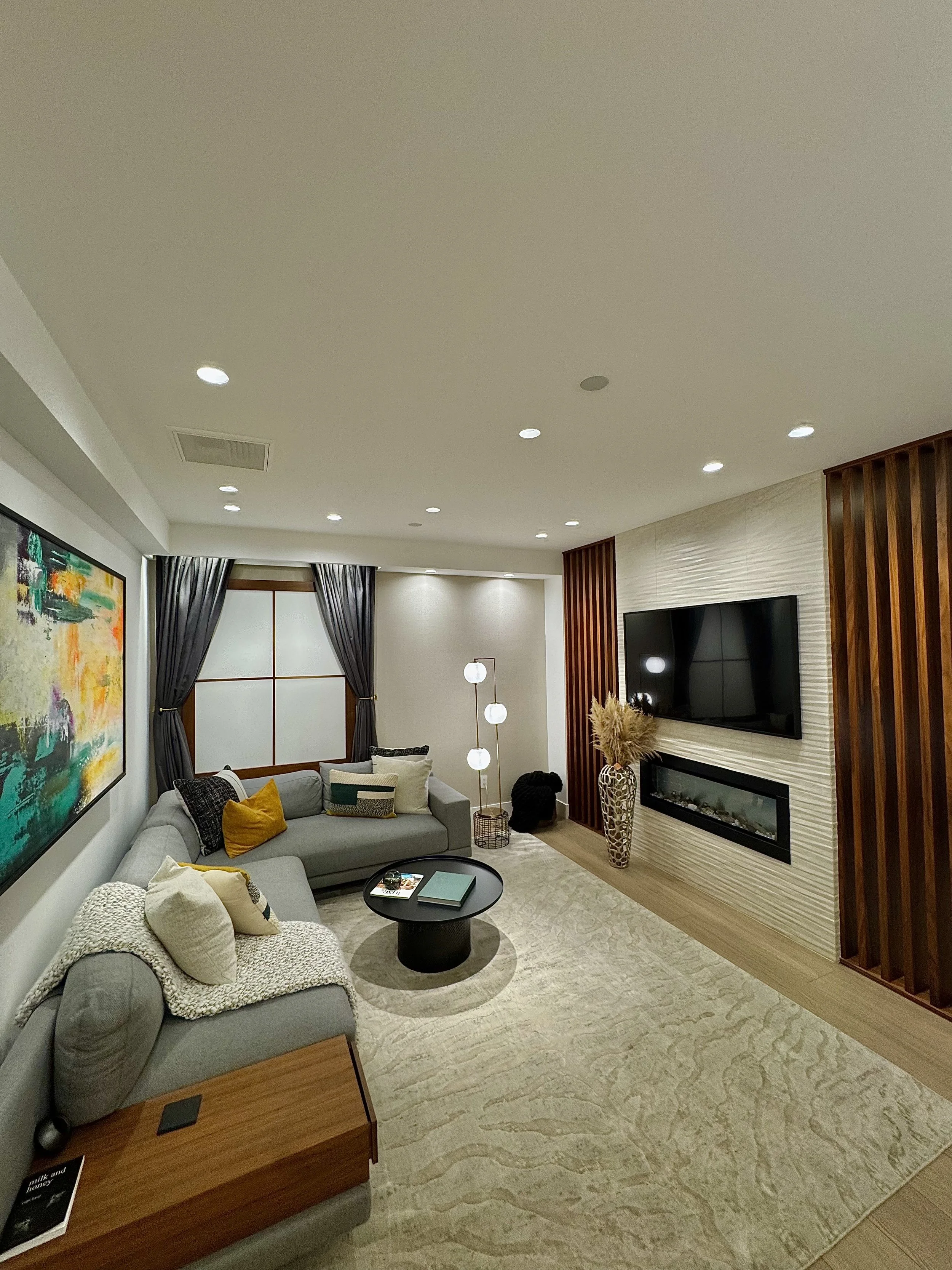 Modern living room with gray sofas, colorful cushions, a black round coffee table, large window with dark curtains, abstract wall art, wooden accents, flat-screen TV, fireplace, and a beige rug.