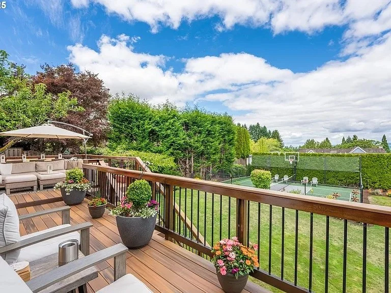 A wooden deck with outdoor furniture and potted plants overlooking a backyard with a basketball court, tennis courts, green grass, trees, and blue sky with clouds.